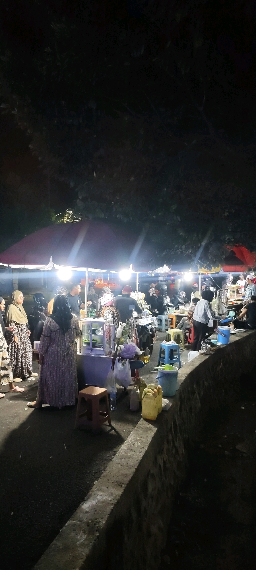 This image depicts a nighttime scene at a street food market in Martapura, Indonesia. The market is set up along a street adjacent to a stone embankment, with trees forming a dark canopy overhead. Several food stalls are visible, illuminated by bright, hanging lights under red and white awnings.
People are gathered around the stalls, some are vendors and others are customers. Several individuals are wearing traditional Muslim attire, including headscarves and caps. There are motorcycles parked around the area, suggesting transportation.
Objects in the scene include plastic stools of various colors (red, blue, yellow), buckets, and containers of what appear to be cooking oil. Plastic bags are hanging from stalls, likely containing food items or goods for sale. One prominent stall features a glass display case.
The overall atmosphere is lively, with a focus on food vendors and a gathering of people engaging in commerce and social interaction in an outdoor setting at night.