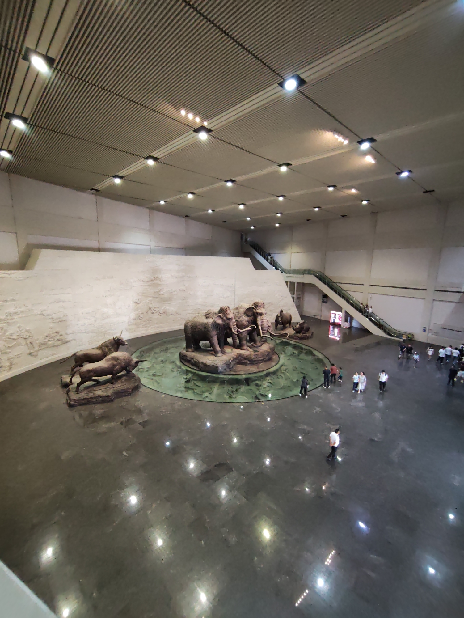 The image shows an indoor scene with a high, slatted ceiling and a polished dark floor. In the center, a circular shallow pool with a green, textured surface surrounds a sculpture of several elephants and other animals, appearing to be a diorama. To the left of the pool, there is a textured wall with bas-relief carvings. An escalator is visible in the background, with people ascending. Several individuals and groups of people are scattered throughout the space, observing the exhibits or walking. The environment appears to be a museum or exhibition hall. The location is identified as Longfeng District, China.