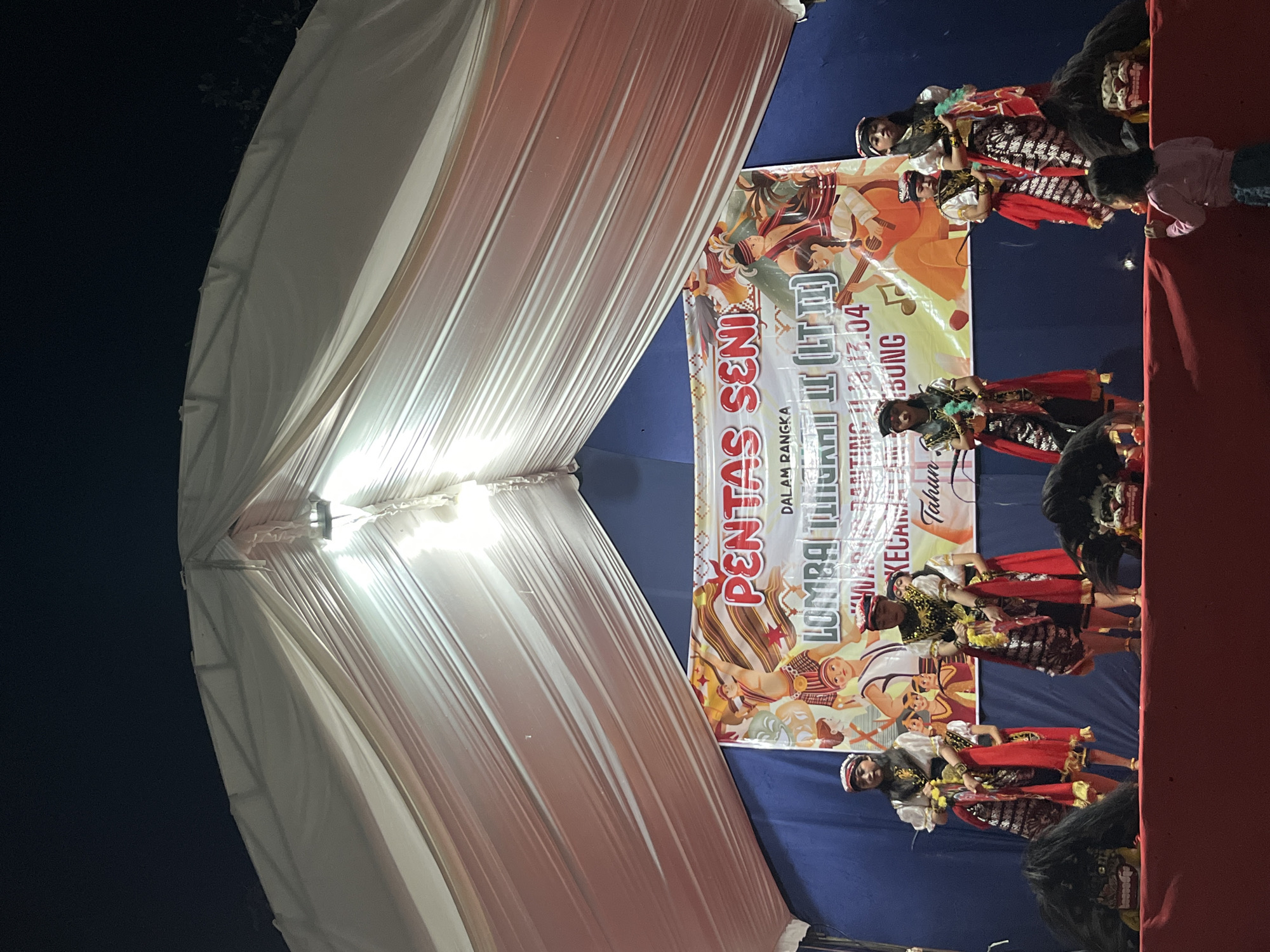 An outdoor stage at night in Gembong, Indonesia, is illuminated by a large white tent overhead and a bright light source. The tent drapes down, creating a backdrop for a cultural performance. In the background, a banner reads "PENTAS SENI" (Art Performance) and "LOMBA TINGKAT II (LT. II)" (Level II Competition), indicating the event's nature. The banner also features colorful illustrations of people in traditional attire. On the stage, six individuals, likely performers, are dressed in elaborate traditional Indonesian costumes, featuring ornate headwear, embroidered tops, and patterned skirts. Two of the performers on the left are facing forward, while the other four are more spread out. In front of the performers, two large, fierce-looking mask-like props, possibly part of the performance, are positioned. A young child in a pink shirt is visible in the foreground, looking towards the stage, suggesting an audience member. The overall scene captures a vibrant cultural event taking place in the evening.
