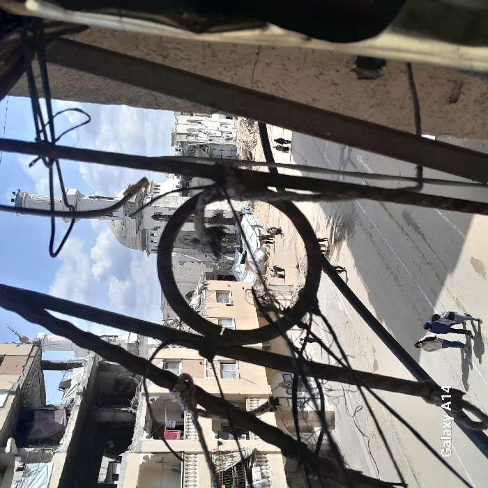 The scene, captured from an elevated perspective through a foregrounded, out-of-focus metal grate, depicts a heavily damaged urban environment in Khan Yunis, Palestinian Territory. A prominent white mosque, featuring a dome and a tall, slender minaret, stands in the mid-background, showing damage to its lower sections and surrounding structures. To the left, multiple multi-story buildings are extensively damaged, with collapsed sections, exposed interiors, and debris; laundry is visible hanging from one damaged balcony. A wide, debris-strewn street runs across the midground. Approximately ten individuals are visible: three people are conversing on the street in the immediate midground, while around seven others are scattered in smaller groups near the mosque and among the rubble, some near improvised tarp shelters. The ground near the mosque consists primarily of dust and construction debris. The sky above is partially cloudy.