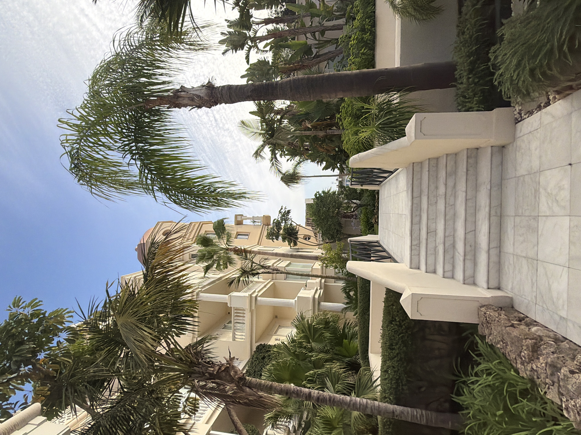 The image depicts a pathway leading up a marble staircase towards a building with balconies and palm trees. The building is a cream-colored structure with white railings and columns. A domed tower is visible on the right side of the building. The pathway is paved with white marble tiles, and the staircase is also made of marble. Black metal railings line the sides of the staircase. Lush green vegetation, including various types of palm trees and bushes, surrounds the pathway and staircase. The sky is blue with scattered white clouds. The scene appears to be set in a resort or a residential complex, possibly in a warm climate. The location is identified as Resinera-voladilla, Spain.