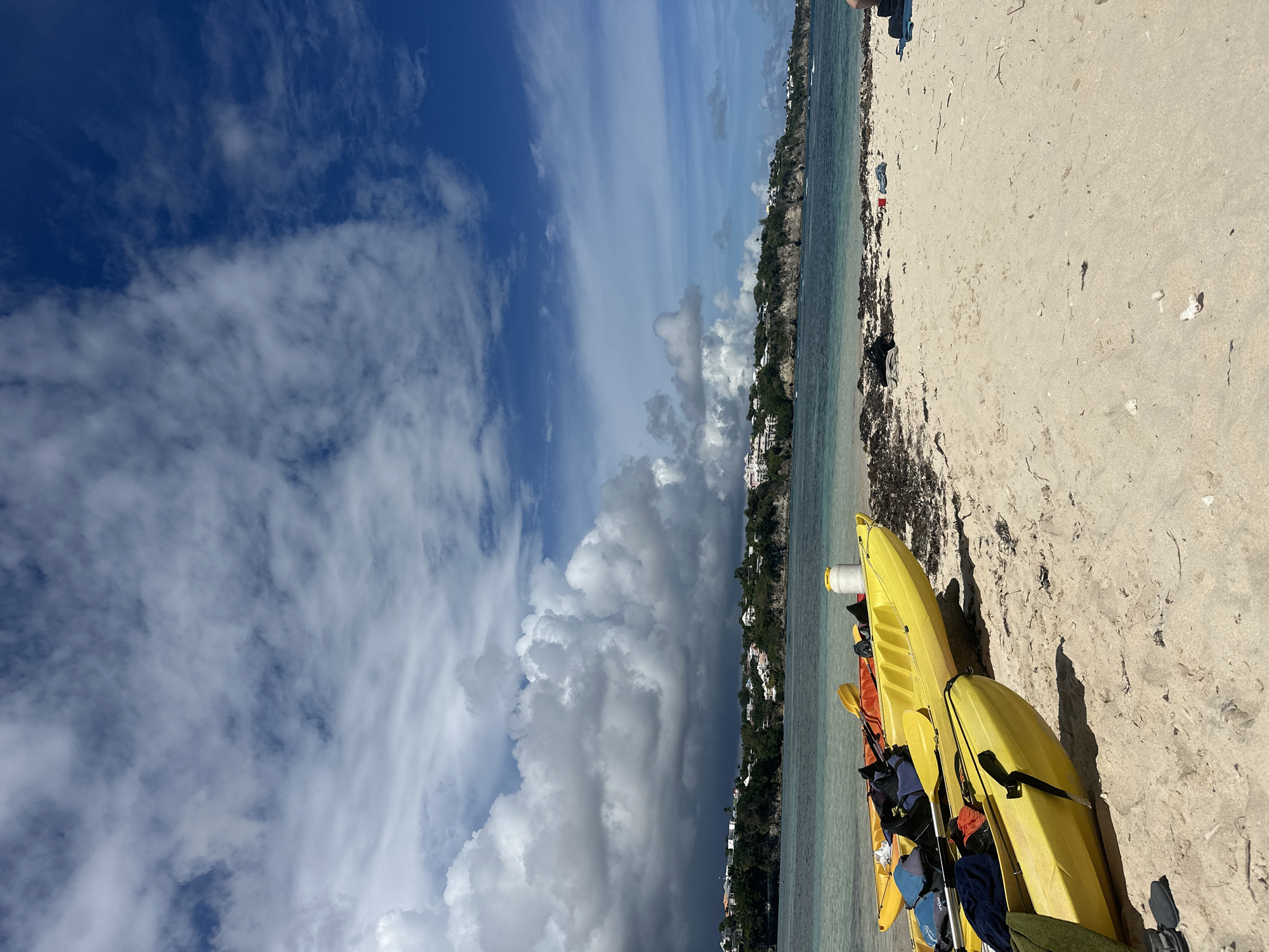 The image shows a beach scene in Le Gosier, France. The perspective is rotated 90 degrees clockwise. On the right side of the image, there is a sandy beach with seaweed. Several yellow kayaks are stacked on the sand near the water's edge. Some paddles and gear are visible in and around the kayaks. The water is clear and calm. Beyond the water is a coastline with buildings and lush vegetation. The sky is predominantly blue with white and gray cumulus clouds.