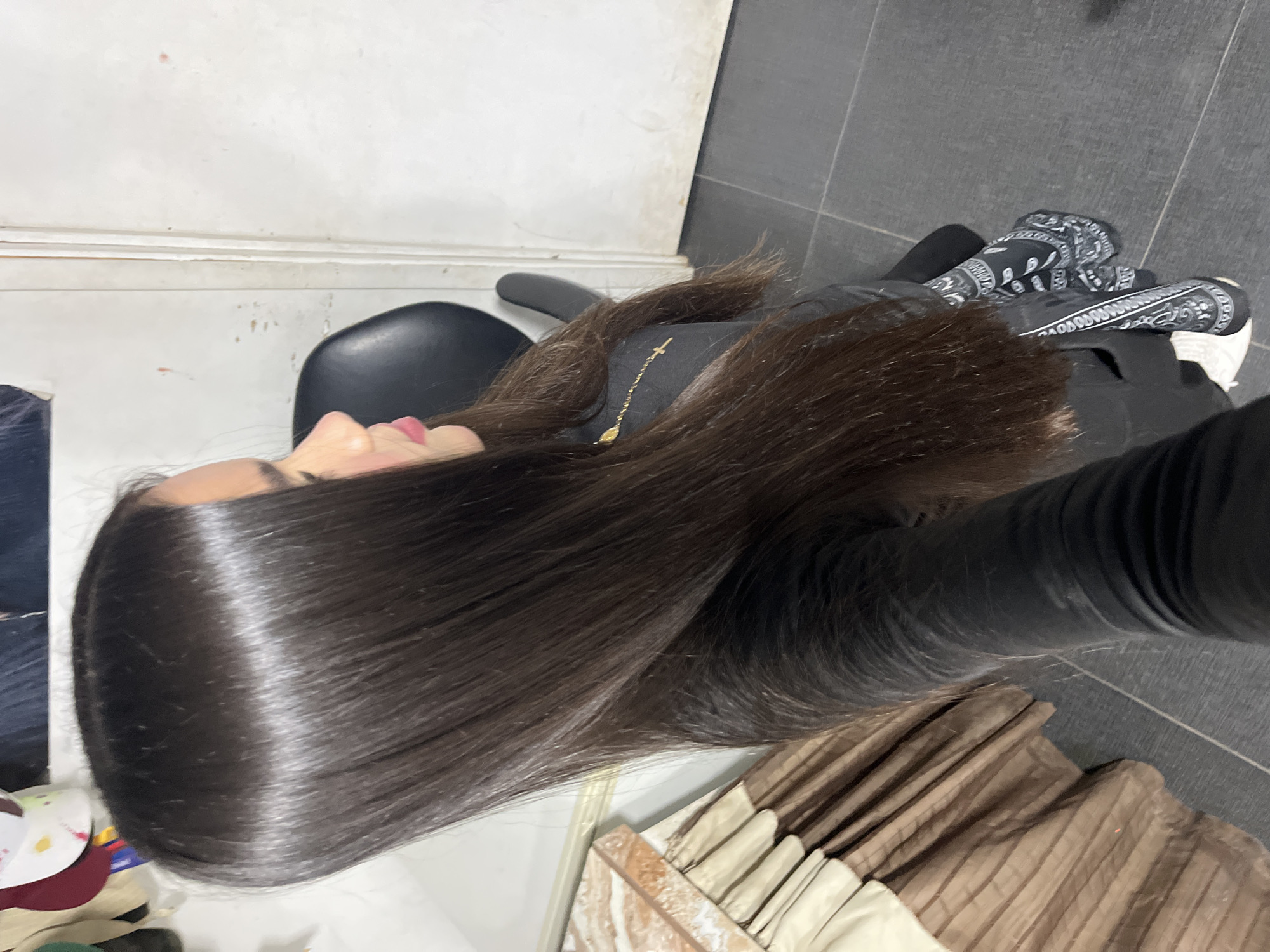 In the city of Juan Griego, Venezuela, a person identified as female is depicted in an indoor setting. The subject, viewed from the upper body in a side profile, possesses long, straight, dark brown hair that appears exceptionally smooth and lustrous, parted near the center and extending well past her shoulders. Her face, seen in profile, exhibits fair skin and pink-tinted lips. She is wearing a long-sleeved black garment. A delicate gold-colored chain with a small cross pendant is visible on her left shoulder. Her left forearm is adorned with a black and white paisley-patterned fabric, possibly a bandana or part of the garment's design. Partially visible on her feet are white athletic shoes.
The subject is seated in a black, high-backed chair, a portion of which is visible behind her. To the left of the frame, a pleated fabric in shades of beige and brown, resembling a curtain or furniture covering, is present. Above this, against a light-colored wall, indistinct objects, possibly headwear, are hanging. The background to the right features a plain white wall with several small, irregular reddish-brown marks or stains. A vertical white door frame is partially visible along the right edge. The floor is composed of dark gray or black tiles.