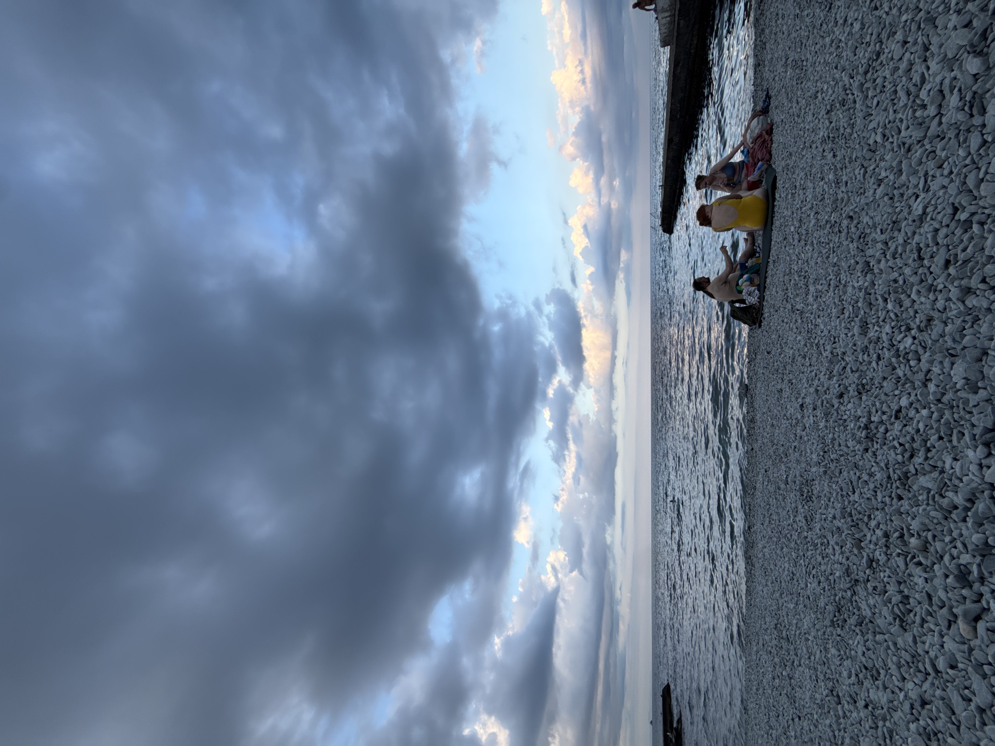A group of three individuals are seated on a pebble beach in Partenit, Ukraine, facing the sea. The sky above is filled with dramatic, dark clouds, with patches of lighter blue and scattered white clouds visible. The sea is calm, with gentle waves. In the distance, a concrete breakwater extends into the water, and a single person is visible sitting on its edge. The beach itself is covered in smooth, grey pebbles. The group on the beach is casually dressed, with one person wearing a yellow swimsuit, another in a patterned swimsuit, and the third in darker clothing. They appear to be relaxing and conversing.