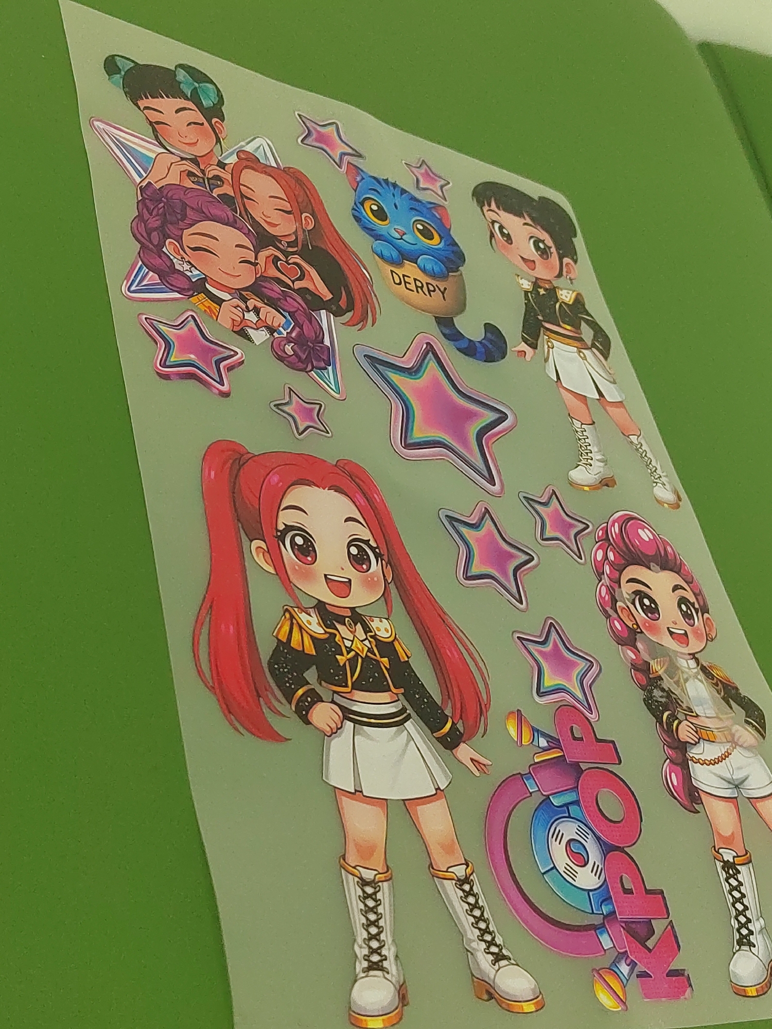 The image shows a sheet of stickers. The stickers depict cartoon-style figures resembling members of a K-pop group, stylized stars, and the word "K-POP" integrated into a graphic design resembling a microphone or sound equipment. There are four distinct female characters, each with different hair colors and hairstyles, wearing outfits that appear to be stylized military-inspired or stage costumes. Another sticker is a blue cartoon cat in a cup with the word "DERPY" next to it.
The sheet background appears to be translucent and is placed on a green surface.
There is no location information such as Votkinsk or Russia present in the image.