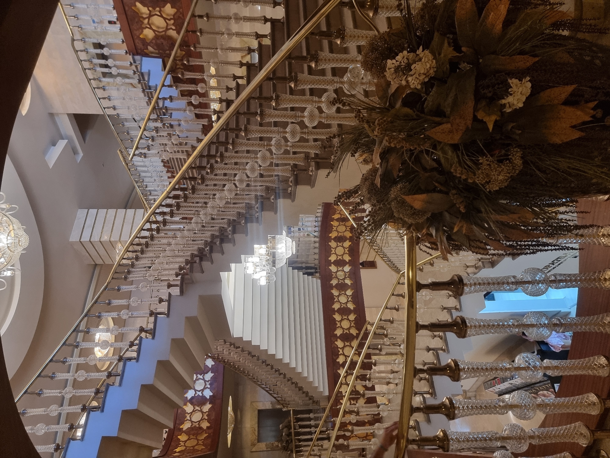 A grand staircase with ornate brass railings and crystal balusters is shown in a hotel lobby. The staircase has multiple landings and curves upwards, with a large floral arrangement in the foreground. The walls are decorated with patterned panels and there are chandeliers on the ceiling. The location is the interior of a hotel in Manavgat, Turkey.
