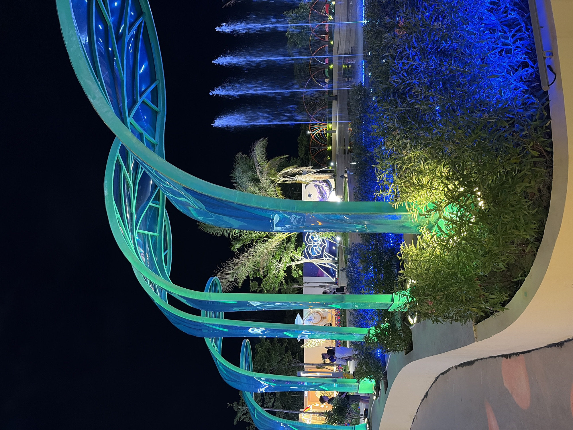 This is a night shot of a public park in Da Nang, Vietnam, possibly the "Dreamy Garden" or a similar illuminated attraction. The foreground features a curved walkway with plants illuminated by blue and green lights. In the mid-ground, several large, translucent, arched structures made of blue and green material, resembling leaves or waves, are lit from within and also cast colored light onto the surrounding foliage. Behind these structures, a fountain sprays water into the air, illuminated with blue light. Palm trees and other greenery are visible, along with people in casual attire, some standing and observing the fountain. In the background, there are various illuminated art installations and structures, suggesting a festive or exhibition-like atmosphere. The overall scene is dark, with the artificial lighting creating a vibrant and somewhat futuristic ambiance.