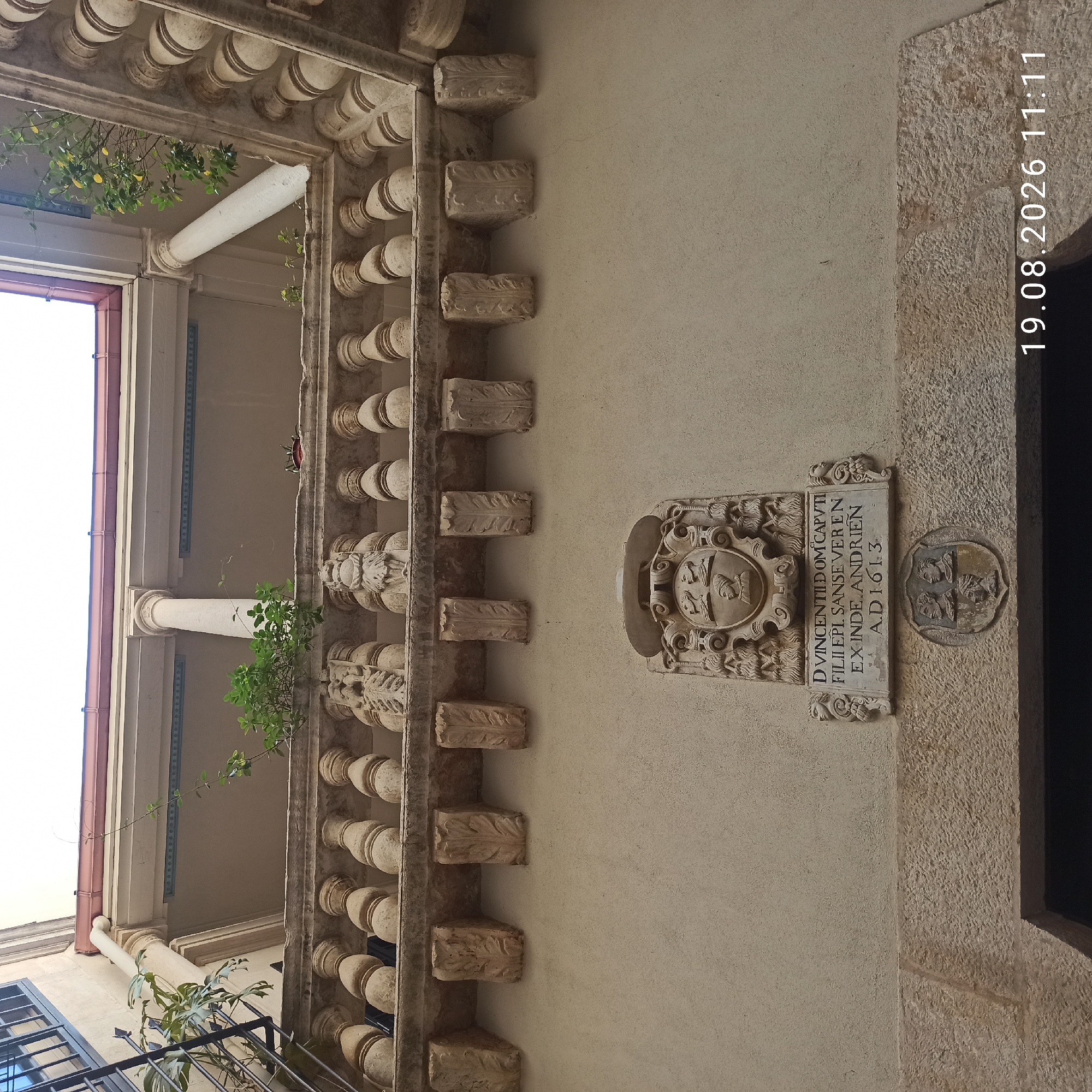 The image shows a close-up, upward view of a stone facade on a building. In the foreground, there is an ornate stone crest with carved profiles of three heads above a shield. Below the crest, a stone plaque displays carved Latin text and the date "A.D. 1613". The facade is made of roughly textured plaster. Above the plaque, a stone balustrade with decorative carvings and balusters runs horizontally across the image. Behind the balustrade, columns and a roofline are visible, suggesting an upper level or courtyard. Green plants are growing on the balustrade and climbing up the facade. The bottom right corner of the image contains a timestamp: "19.08.2026 11:11". The architecture suggests a historic building, possibly in Southern Italy. This is the former Palazzo Stola (now Palazzo Baronale) in Ruvo di Puglia, Italy, specifically the entrance portal featuring the family crest of Vincenzo Domini and the date related to a construction or renovation.