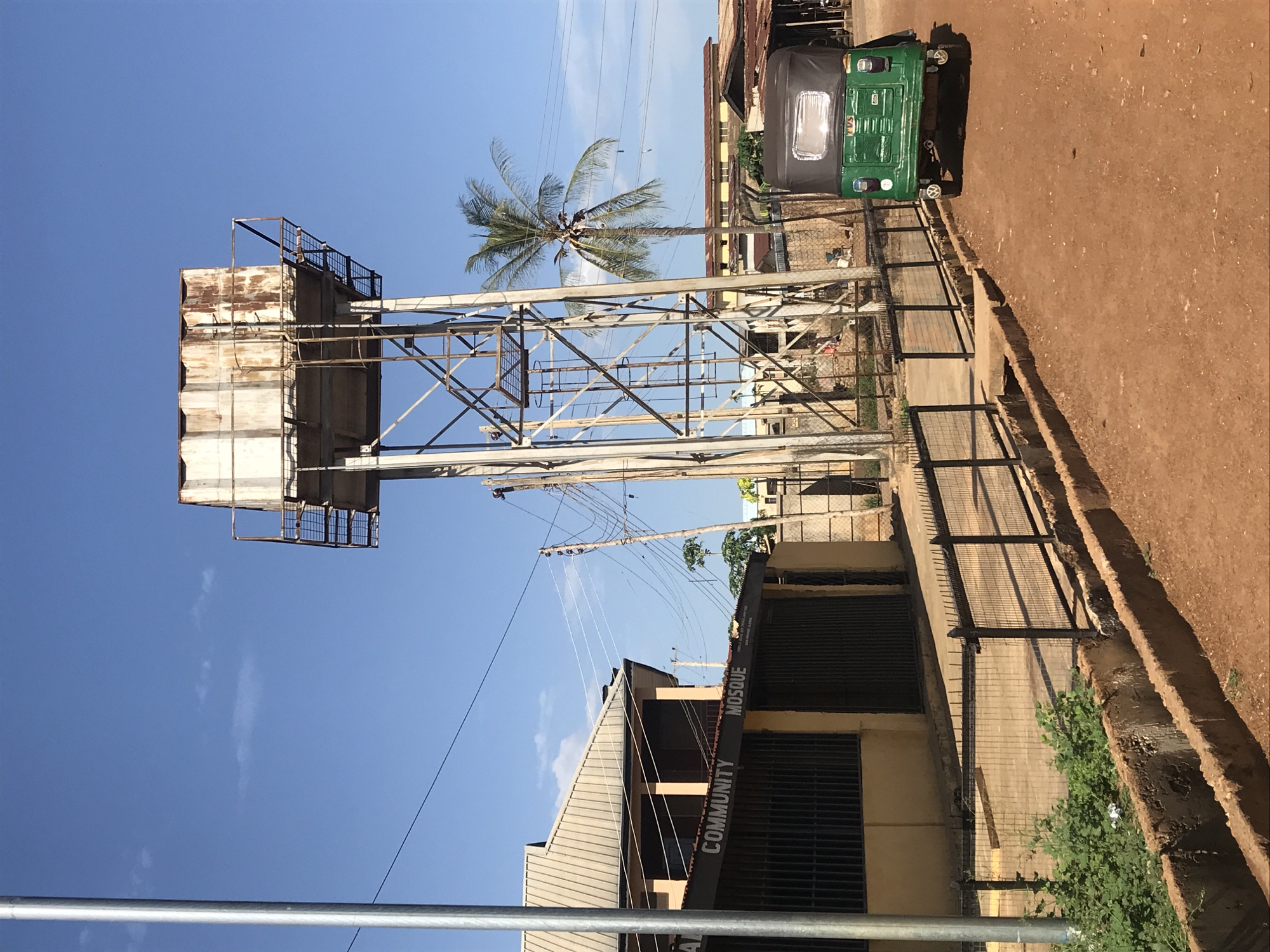 The image presents a sunlit outdoor scene in Mandate Estate, Ilorin, Nigeria, under a clear blue sky dotted with a few faint wispy clouds. The photograph is oriented vertically, showing the ground extending from the bottom-right towards the top.
The dominant feature is a tall, metal-framed water tower situated prominently in the left-center. It supports a large, rectangular water tank that appears weathered, with sections showing discoloration and possibly rust, indicative of age or heavy use. A metal walkway or railing can be seen around the top perimeter of the tank.
Behind the water tower and slightly to its right, a tall, slender palm tree with lush green fronds stands out against the sky. A network of overhead utility poles and power lines stretches across the mid-ground, leading towards a building in the lower-left portion of the frame. This structure features a corrugated metal roof, light-colored walls, and windows protected by dark metal bars. Clearly visible text on the building's facade reads "COMMUNITY" and "MOSQUE," identifying it as a community mosque.
On the right side of the image, a wide, reddish-brown dirt road or unpaved path is present. A green and black auto-rickshaw, commonly known as a 'Keke Napep' in Nigeria, is parked on this road, displaying "TVS" on its front. Metal fences and railings run along the edges of the building and the dirt path, with some green shrubs growing near the base of the fence.
The scene is static, capturing a slice of daily life in a bustling urban or semi-urban environment. The bright illumination suggests it is mid-morning or afternoon.