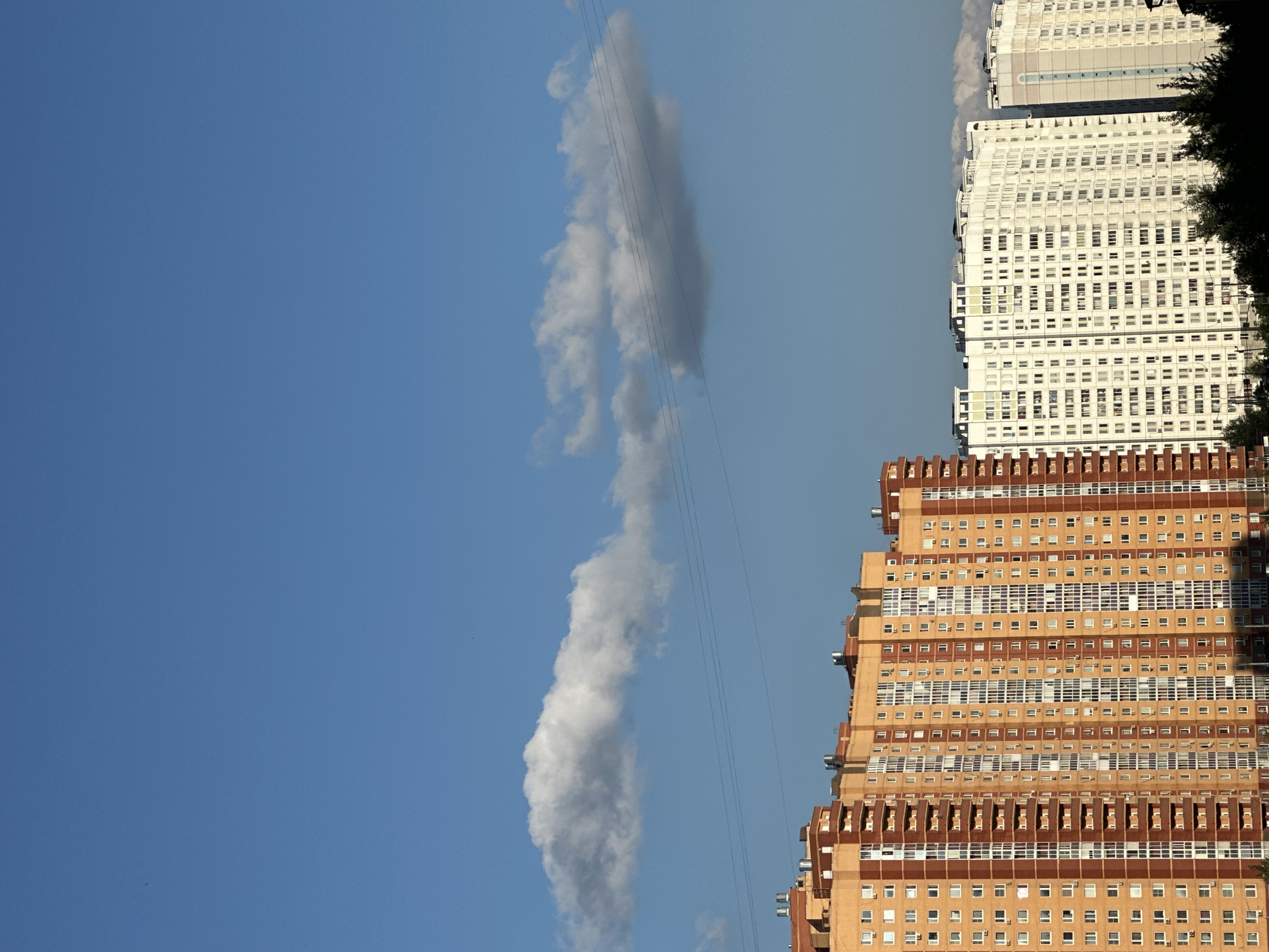 The image depicts a clear blue sky with scattered white clouds and power lines stretching horizontally across the frame. Below the sky are several tall residential buildings. On the left, a large, multi-story apartment building with an orange and brown facade is prominent. To its right, a series of white apartment buildings stand in a row, with one to the far right appearing slightly lighter in color. The buildings have many windows and balconies. At the bottom of the image, the dark silhouettes of trees and other foliage are visible, suggesting a natural element within the urban landscape. The scene is likely a view of a residential area in Podolsk, Russia, characterized by its modern high-rise apartment complexes against a backdrop of the sky.