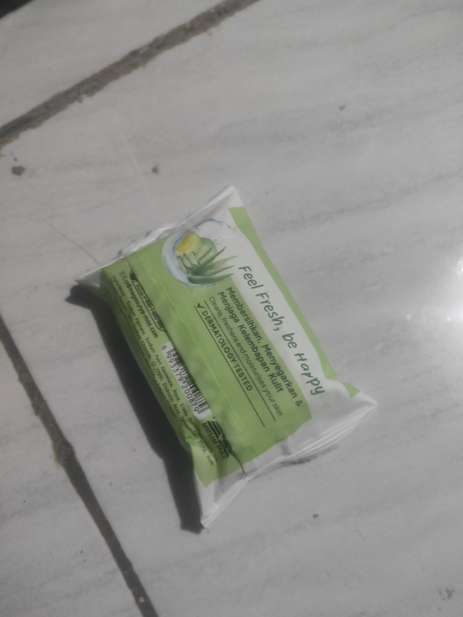 A green and white soap wrapper is lying on a tiled floor. The wrapper is oriented diagonally across the image, with the top left corner of the wrapper closest to the viewer. The wrapper has text printed on it in both English and Indonesian, along with a barcode and a picture of aloe vera. The tiles on the floor are a light gray color and appear to be weathered. There are shadows cast on the floor, indicating that there is a light source from the left. The scene is indoors, and the overall lighting is dim. There are no people or animals visible in the image. The location is in Antirogo, Indonesia, but this is not evident from the image itself.