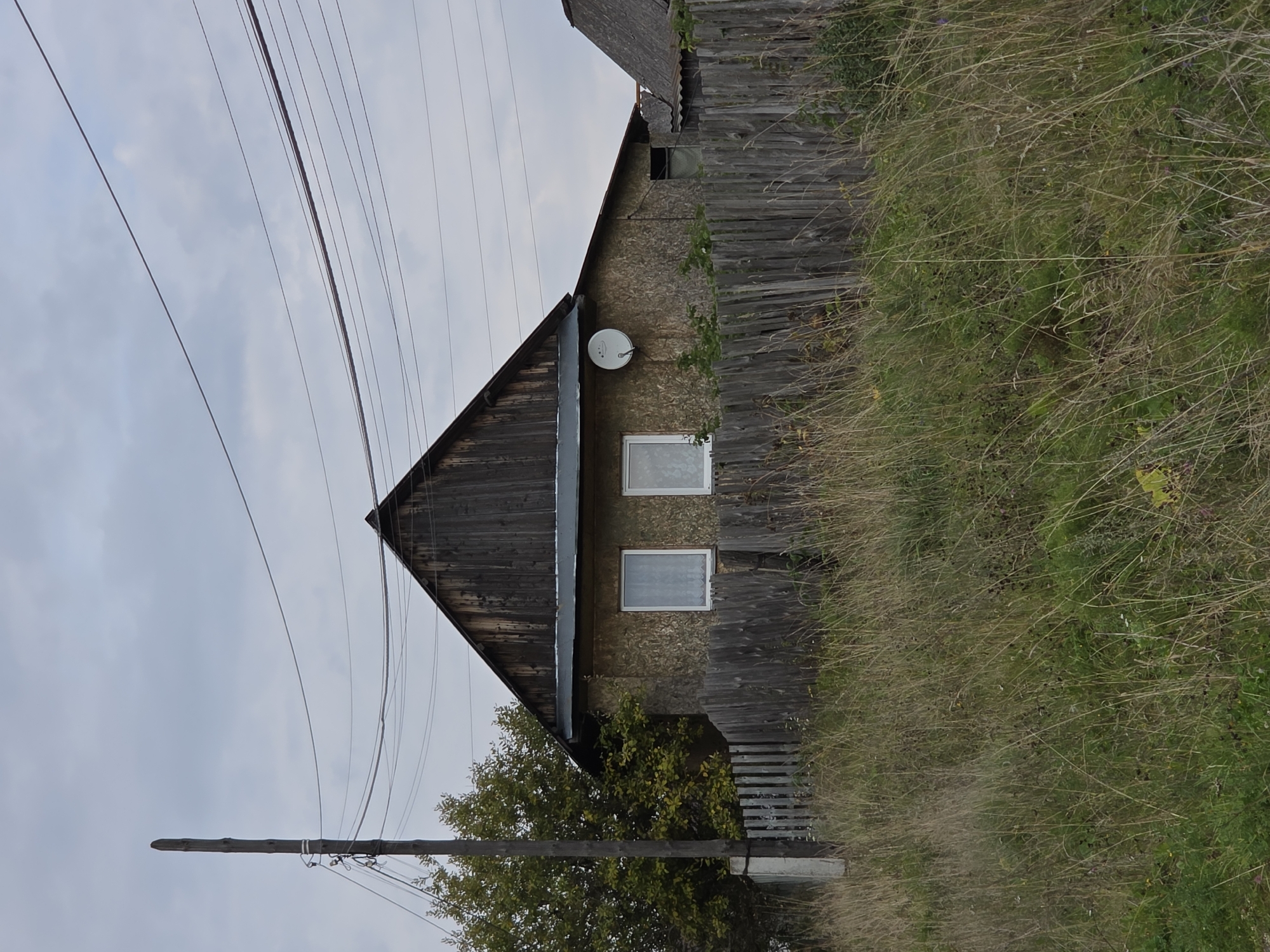 This is a forensic, matter-of-fact description of an image depicting a rural scene. The image shows a wooden house with a dark wooden gable roof. The walls of the house are covered with a textured material, possibly oriented strand board (OSB). There are two windows on the front of the house, each with white frames and curtains. A satellite dish is mounted on the right side of the house. A weathered wooden fence runs in front of the house. The area in front of the fence is overgrown with tall, dry grass and some scattered green vegetation. To the left of the house, there is a tall wooden utility pole with multiple overhead wires attached. Behind the house, on the left, is a dense tree with green and yellow foliage. The sky is overcast with a gray, cloudy appearance. The image appears to have been taken outdoors in a rural or semi-rural setting, possibly in or near Magnitka, Russia, given the context provided. There are no people or animals visible in the image.