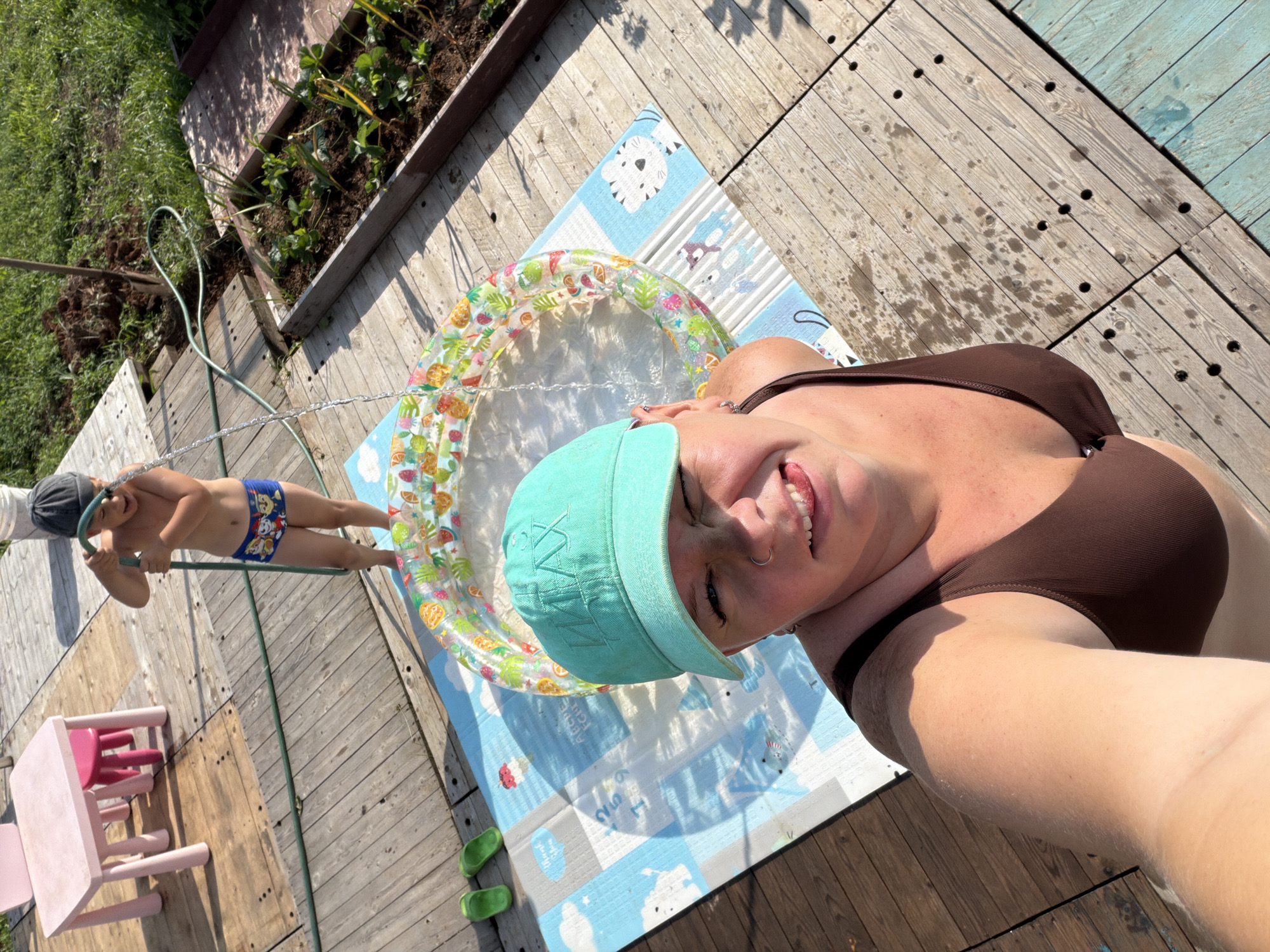 A child wearing blue Paw Patrol swimming trunks and a dark gray beanie is spraying water from a green hose into an inflatable kiddie pool. The pool is located on a blue and white mat. A woman wearing a brown swimsuit and a teal baseball cap is taking a selfie, sticking her tongue out. She has piercings in her nose and ear. In the background, there are two pink children's stools and a pink table, as well as a raised garden bed with plants. The scene takes place on a wooden deck on a sunny day. The location is a backyard in Chaikovsky, Russia.