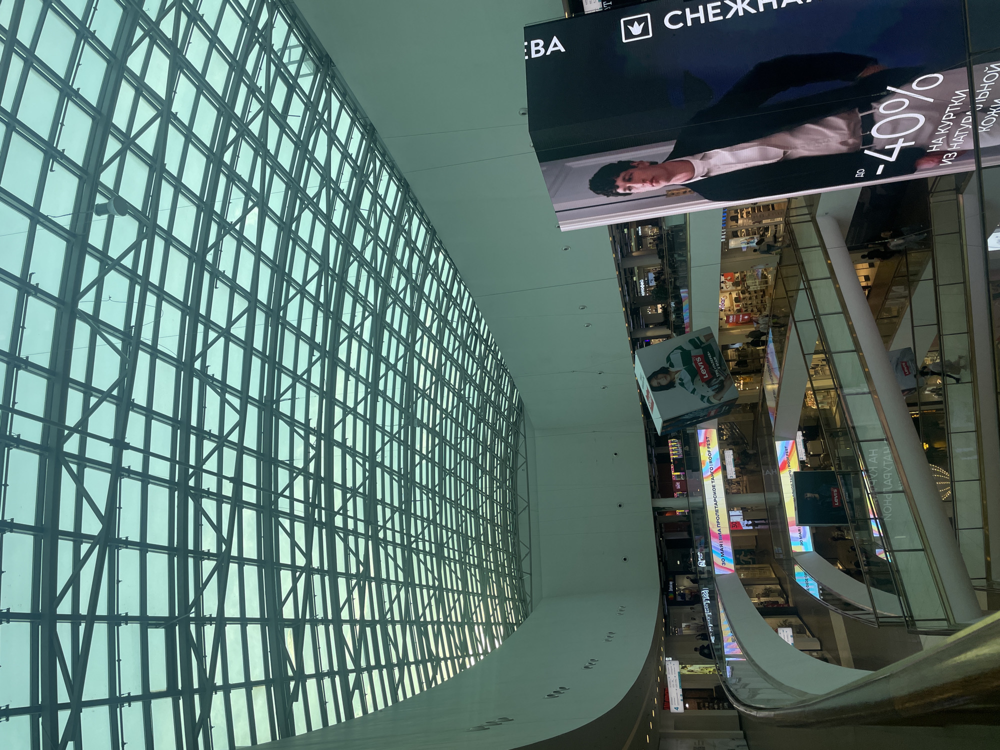The image shows the interior of a multi-level shopping mall in an unknown city. A large glass grid window dominates the left side of the frame, filtering the outside light. The interior space features a multi-level design with escalators, walkways, and various retail stores visible. Digital displays and billboards are present, advertising brands such as Levi's and featuring sales promotions like "-40% on leather jackets". The angle of the shot and the architectural design suggest a modern, high-end shopping environment.