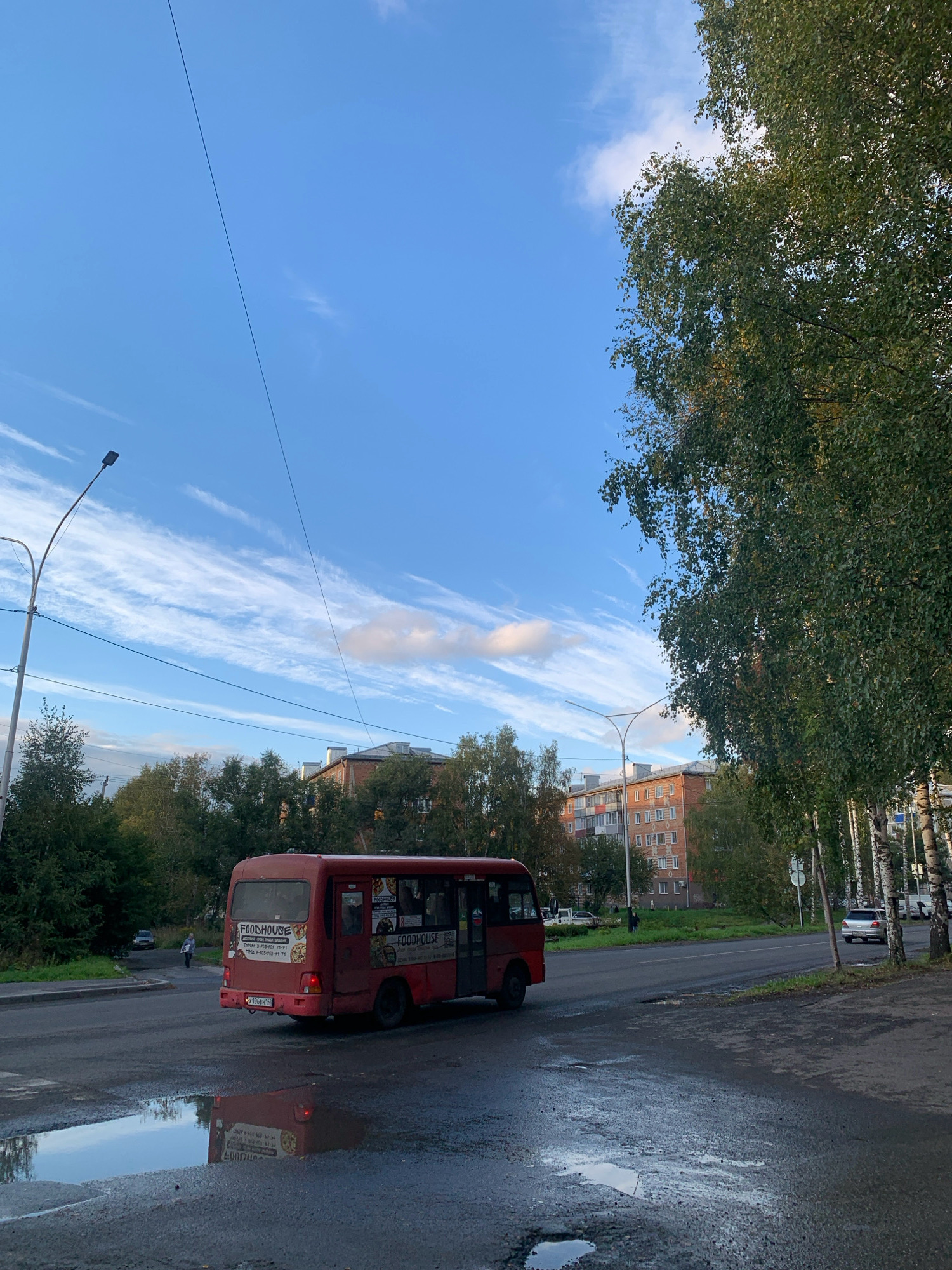 The image depicts a street scene in Prokopyevsk, Russia, under a sky with scattered white clouds. A red minibus, labeled "FOODHOUSE," is traveling on the road. The road surface is wet, with several puddles reflecting the sky and the bus. In the background, there are residential buildings and trees. A lamppost is visible on the left side of the road. A person is walking on the sidewalk in the distance. The overall atmosphere suggests a cloudy day in an urban environment.