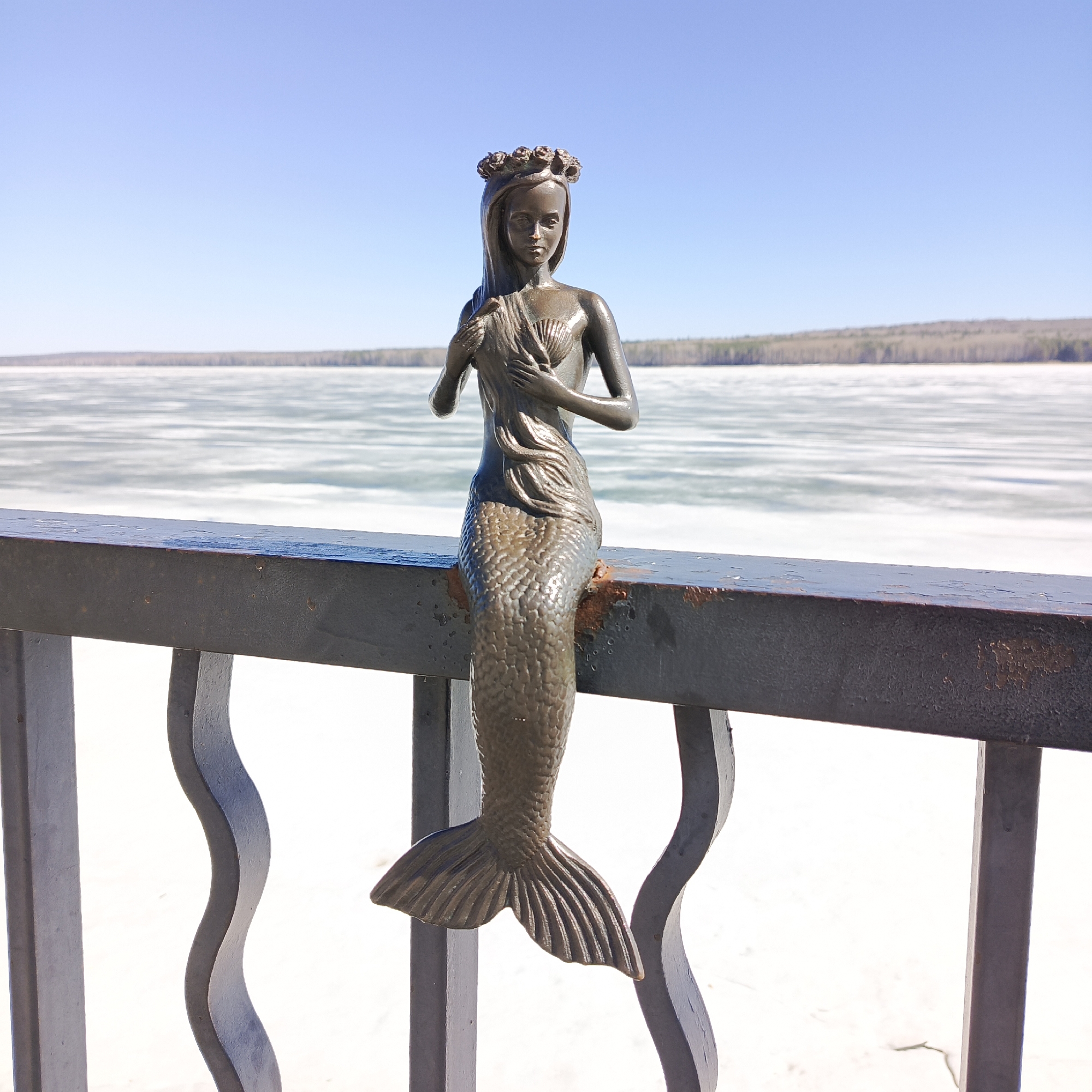 A bronze sculpture of a mermaid sits perched on a dark metal railing. The mermaid has long hair, a flower crown, and her hands are positioned near her chest. The railing is weathered with some rust visible. Behind the railing and the mermaid, a body of water is visible, appearing frozen or covered in ice. In the distance, a line of trees is present under a clear, light blue sky. The sculpture is located in the city of Chaikovsky, Russia.
