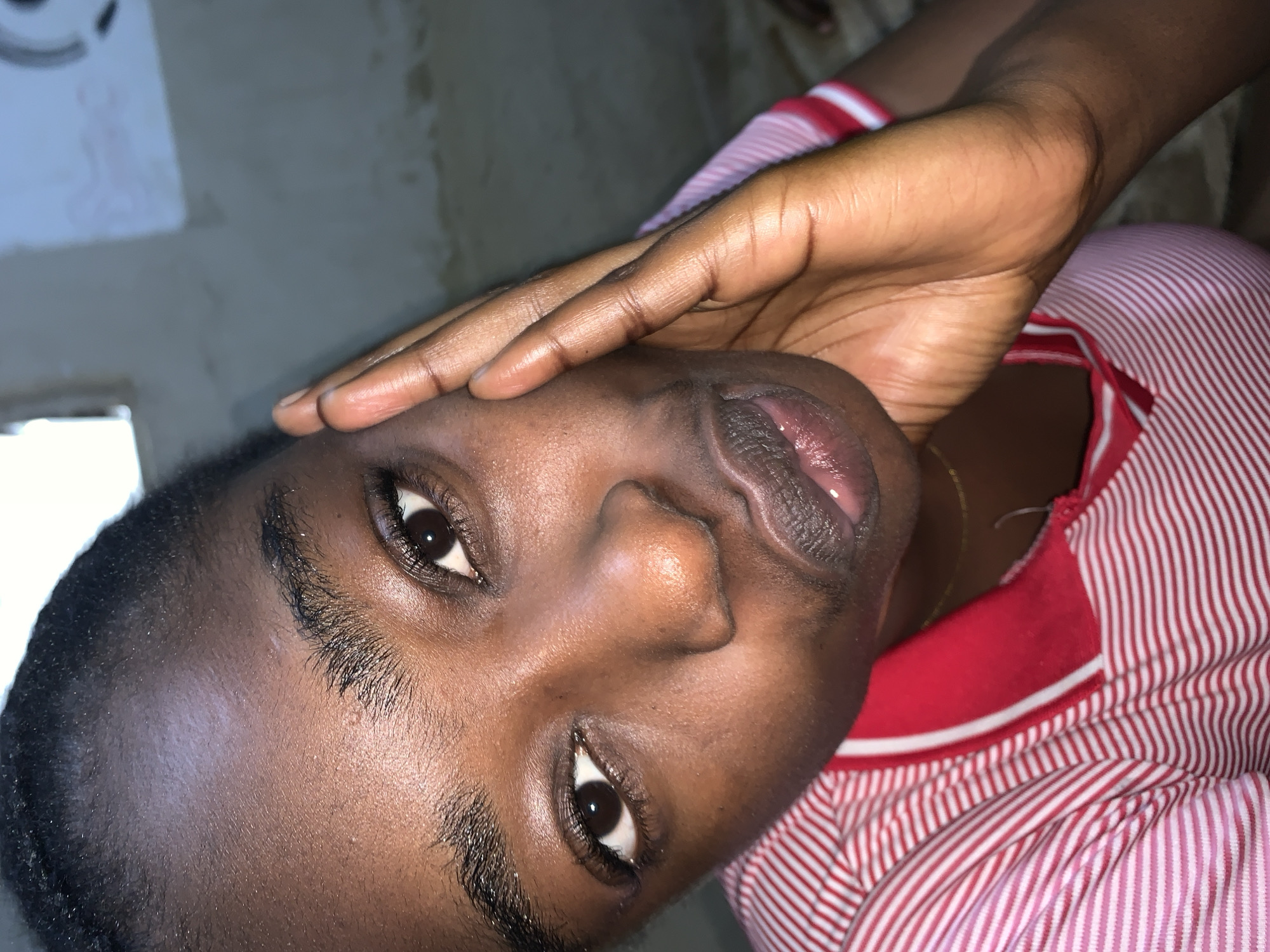 The image is a close-up portrait of a young Black woman. She is looking directly at the camera with her right hand on her cheek. Her lips are slightly parted, and she appears to be wearing lip gloss. She has dark eyes, dark eyebrows, and her hair is pulled back. She is wearing a red and white striped shirt with a red collar. The background is out of focus and appears to be an indoor setting with a plain wall and a window. There is no specific indication of the location being Rafin Par, Nigeria.