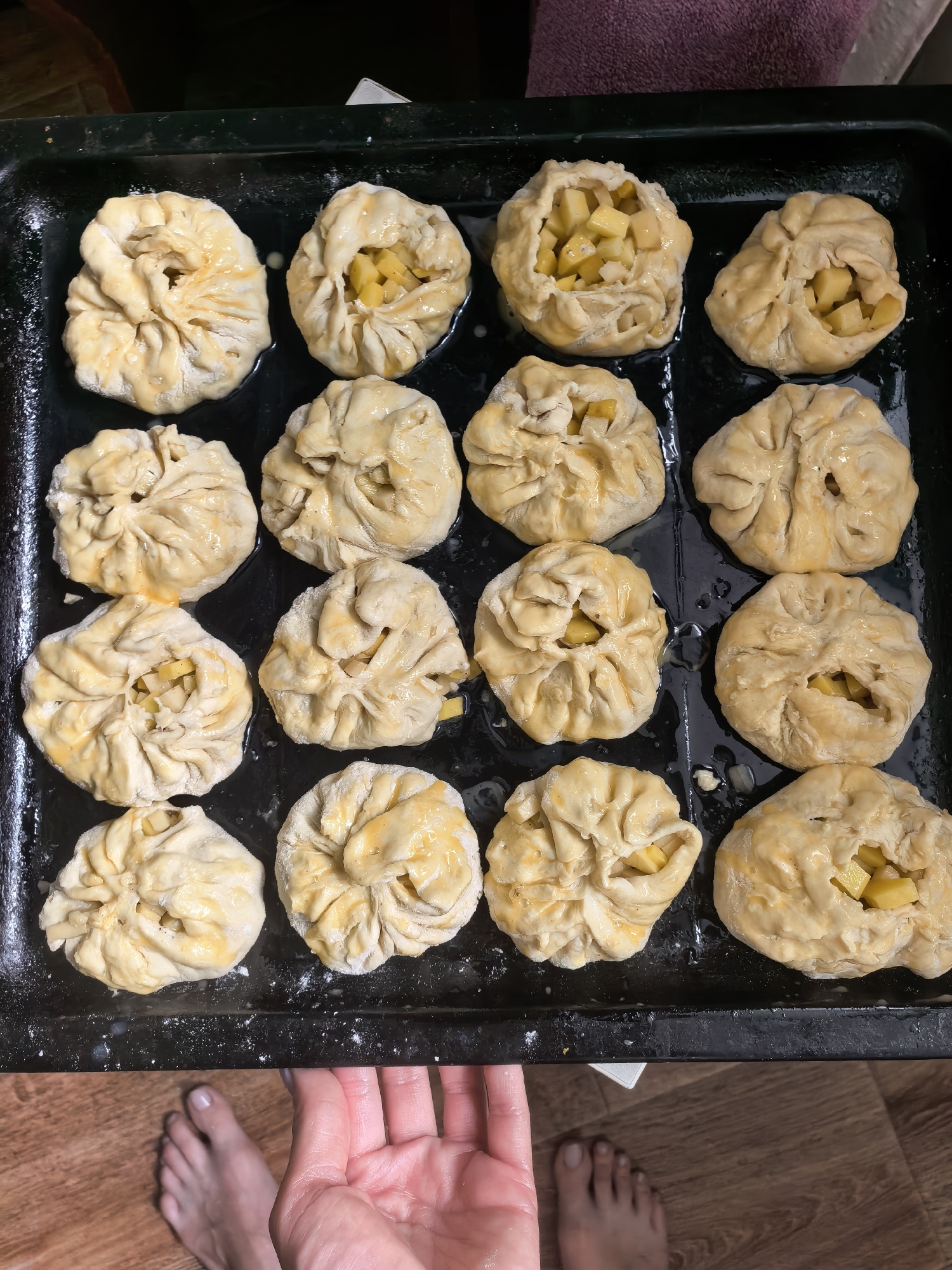 A close-up, high-angle shot shows a black baking sheet filled with sixteen pastry items, possibly uncooked pies or buns. The pastries are arranged in a 4x4 grid. They appear to be made of dough formed into a circular shape with a textured surface and a central opening, some containing diced fruit filling, possibly apples. A clear liquid, likely melted butter or egg wash, is visible on the pastries and in the baking sheet.
In the foreground, a fair-skinned human hand with short nails is visible, slightly blurred. A set of bare human feet, also fair-skinned, are positioned near the bottom left corner.
The location is unknown. There is no information in the image to suggest it is located in the city of Sterlitamak, Russia.