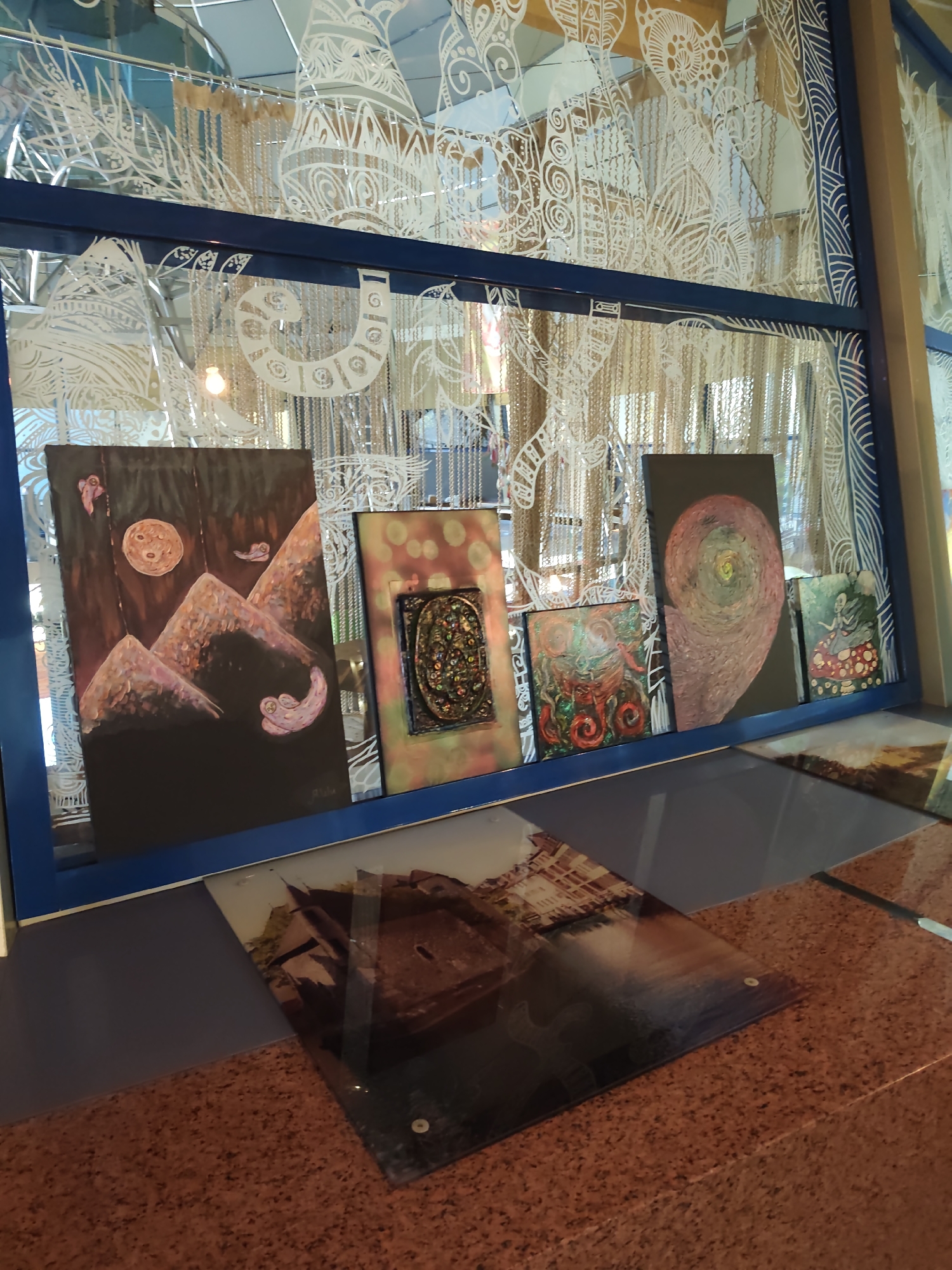 The image shows a display of artworks behind a blue frame. The artworks are various paintings and a textured mosaic. The leftmost painting depicts a surreal landscape with mountains, a moon, and floating figures. To its right is a framed mosaic with intricate details and a variety of colors. Next are two smaller square paintings, one with a red and green abstract design, and another showing a figure in a patterned dress. To the right of these is a larger painting with a swirling, colorful spiral design, and a very small painting on the far right depicting a figure sitting on a mushroom. In the foreground, partially obscuring the artworks, is a glass surface with an image of a canal and buildings, likely Venice. The background appears to be a window or glass partition with decorative white cutouts and beaded curtains. The scene is lit by overhead lighting, creating reflections on the glass surfaces. The location is a display area, possibly an art gallery or a shop. The image does not contain enough information to ascertain its location in the city of Sochi, Russia.