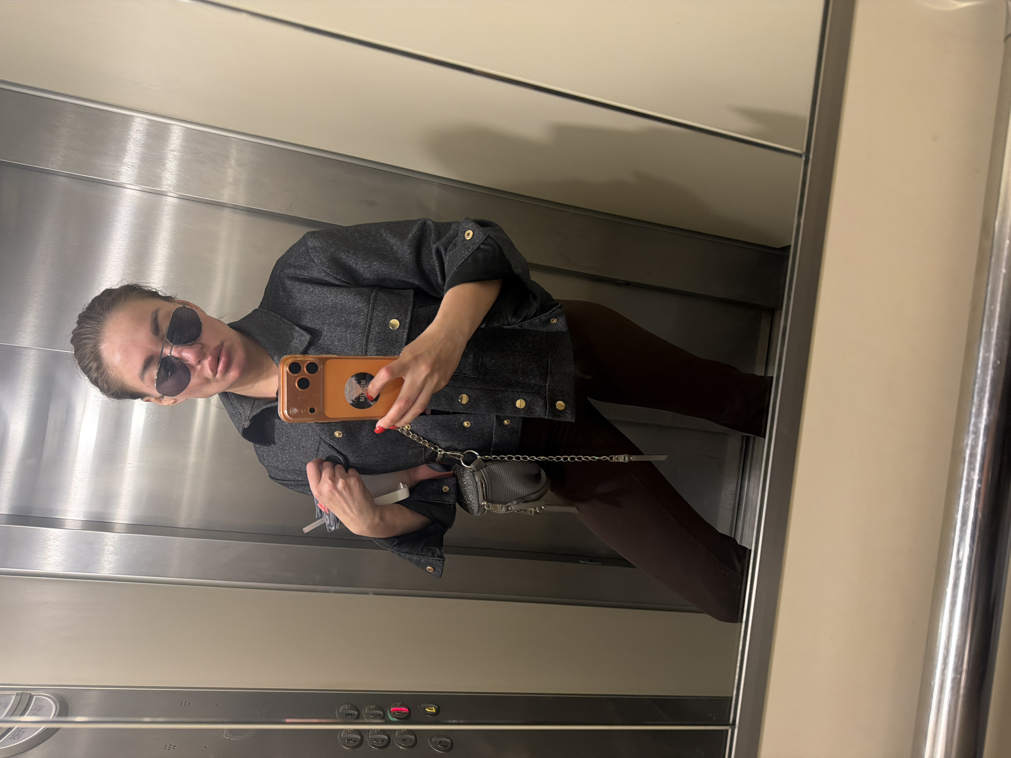 A woman is taking a selfie in what appears to be an elevator. She is wearing sunglasses, a dark gray jacket with gold buttons, and brown pants. She is holding an orange-colored smartphone in her right hand and a small white object (possibly a vaping device) in her left hand. A small, gray purse with a chain strap is visible. The interior of the elevator has stainless steel walls and a panel with buttons. Based on the architectural style of the elevator cabin and the woman's attire, it is unlikely to be in Bar, Montenegro.