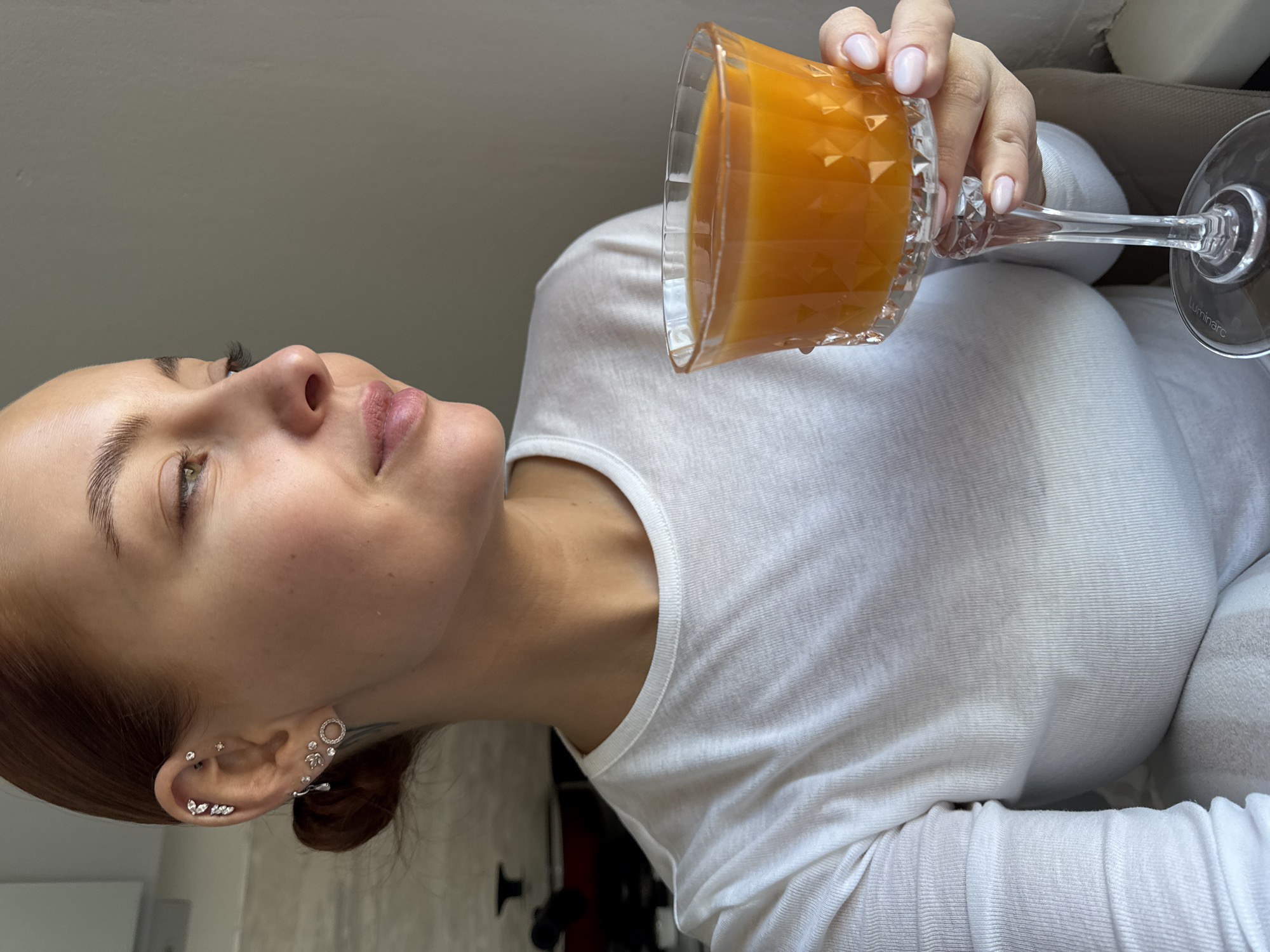 A young woman with auburn hair pulled back in a bun holds a glass of orange juice up to her lips, apparently ready to drink from it. She has fair skin and multiple piercings in her left ear. She is wearing a white long-sleeved shirt. The glass is ornate and tall, with a clear stem. The woman is indoors. The bottom right of the glass is marked with "Luminarc." Although a specific location in the city of Chaikovsky, Russia, cannot be identified from the image, it is presumed to be within a residence or other indoor location.