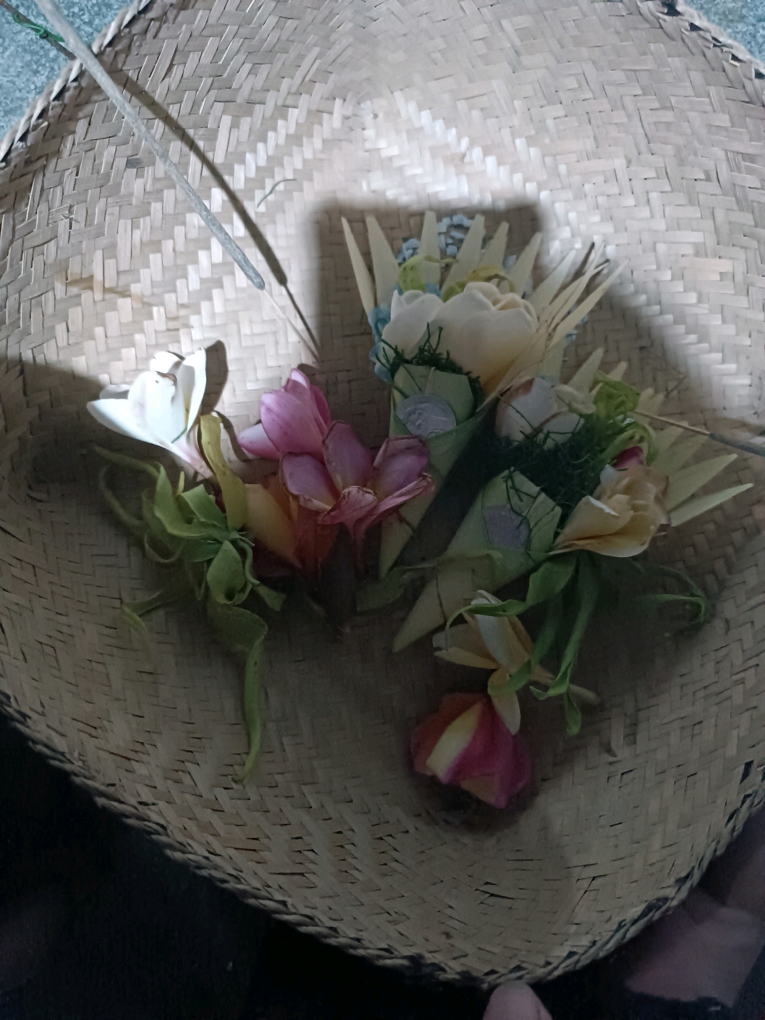 The image depicts a woven basket made of bamboo, likely situated in Kediri, Indonesia. Inside the basket are several offerings, commonly known as "canang sari" in Balinese culture, which are often used in Hindu religious ceremonies. These offerings are typically composed of flowers, rice, and other natural materials, presented in small woven containers. In this specific image, the offerings include a variety of flowers in shades of white, pink, and yellow, along with greenery and what appear to be small pieces of dried palm leaf. Two incense sticks are also present, one resting across the top edge of the basket and another partially submerged within the offerings. The basket is placed on a dark surface, and the lighting suggests it is indoors or under some form of artificial light, casting shadows on the woven material.