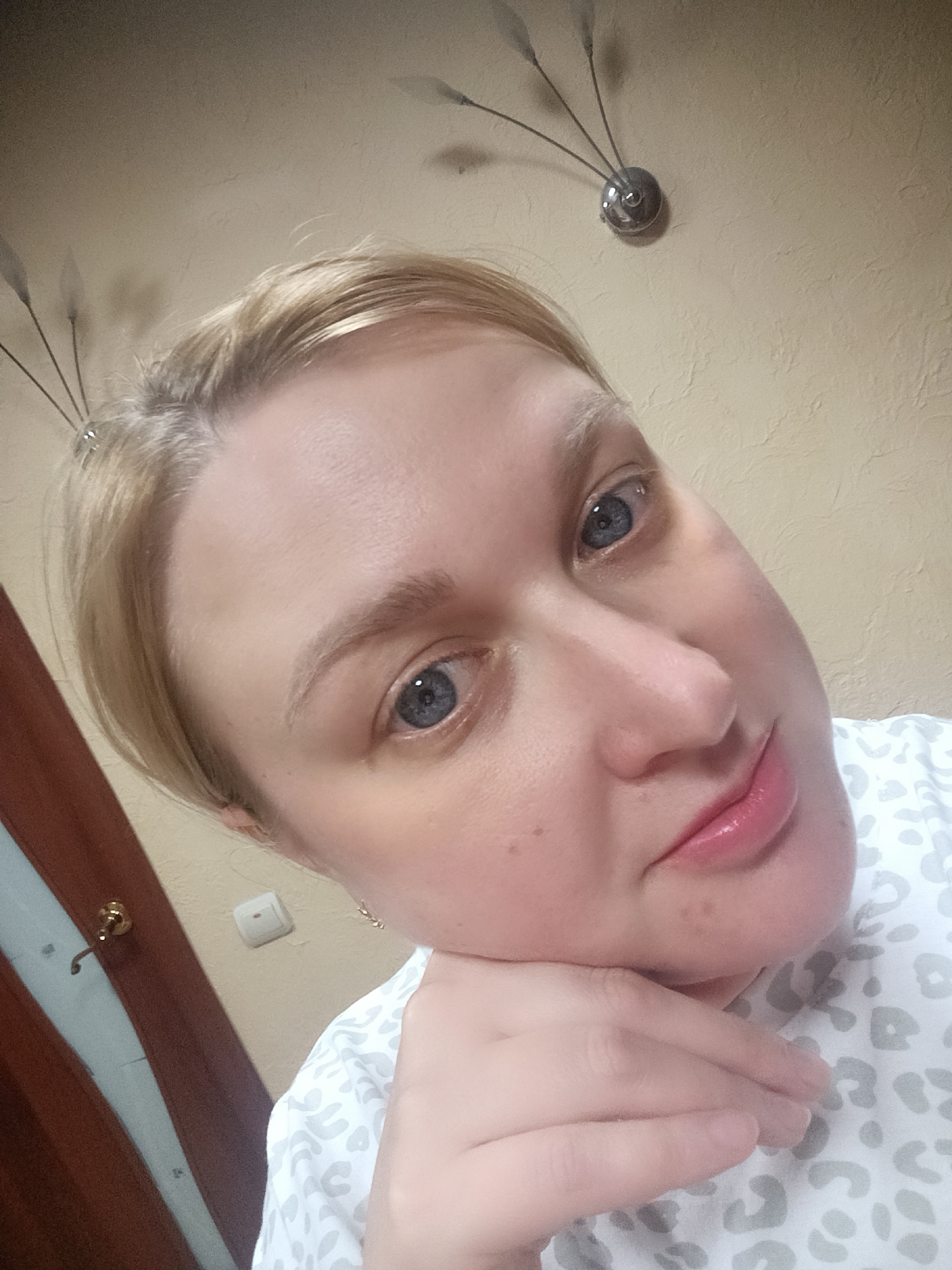 A blonde woman with blue eyes is featured in a close-up, slightly tilted portrait, possibly a selfie. She has fair skin and short, light-colored hair, and is looking directly at the camera with a gentle smile. Her right hand is supporting her chin and jawline. She is wearing a light-colored top with a subtle, diffuse animal print pattern.
The setting appears to be indoors, likely a home in Chaikovsky, Russia. The background wall is a light beige or cream color with a textured finish. In the upper left corner, a decorative wall light fixture with multiple slender arms topped by elongated, translucent elements is visible. To the left, a section of a dark brown wooden door with a brass-colored hook can be seen, along with a white electrical light switch on the wall. The lighting is soft and even, suggesting an indoor environment without strong natural light cues. The woman's expression conveys a sense of calm and approachability. There is no visible text in the image.