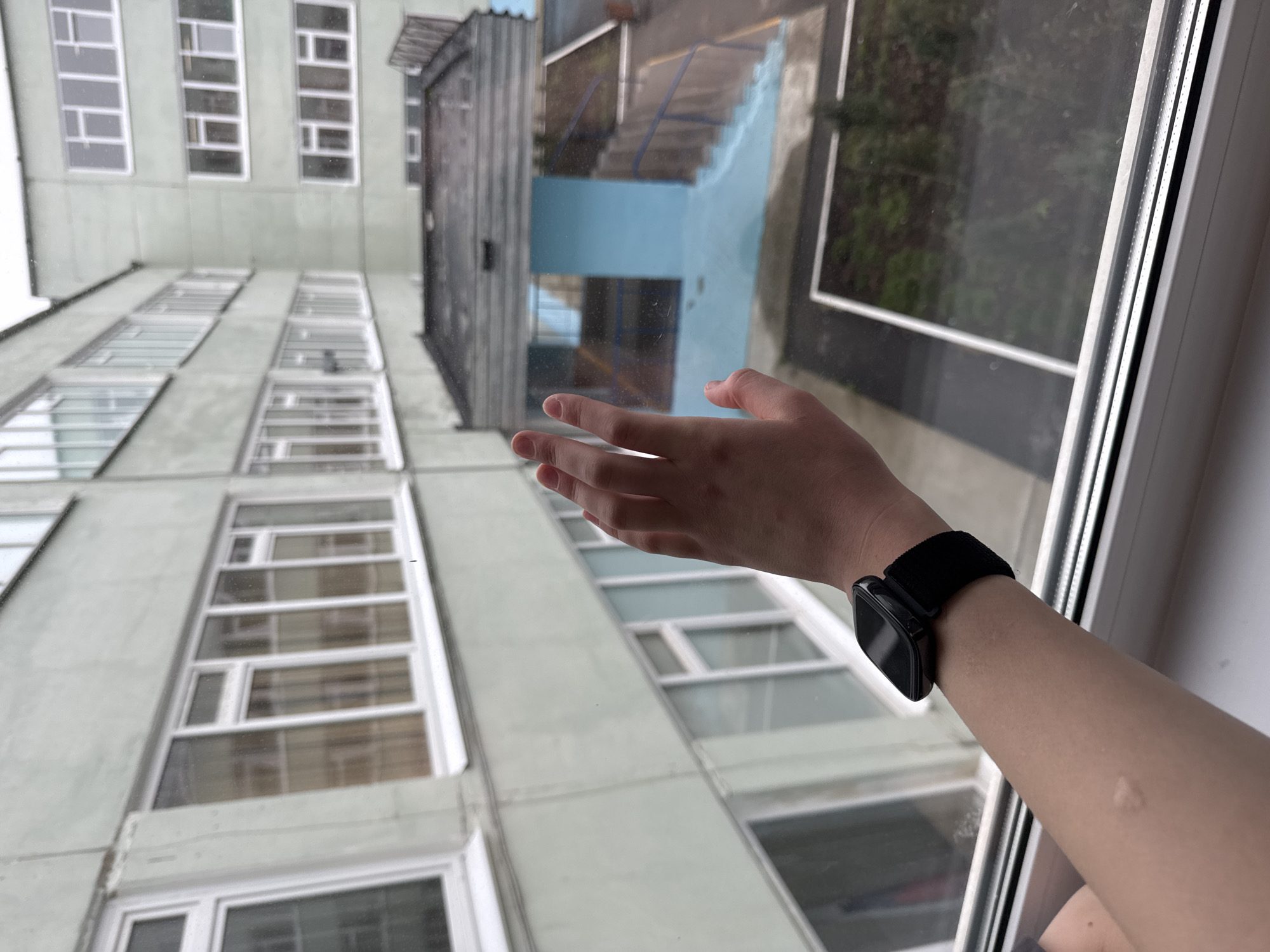 A first-person, upward-angled view through a window shows a hand with a black smartwatch reaching towards the window pane. The window reflects an outdoor staircase painted blue and gray. Beyond the reflection, a multi-story building with numerous windows is visible. The location appears to be in Chaikovsky, Russia, based on the architecture of the building.