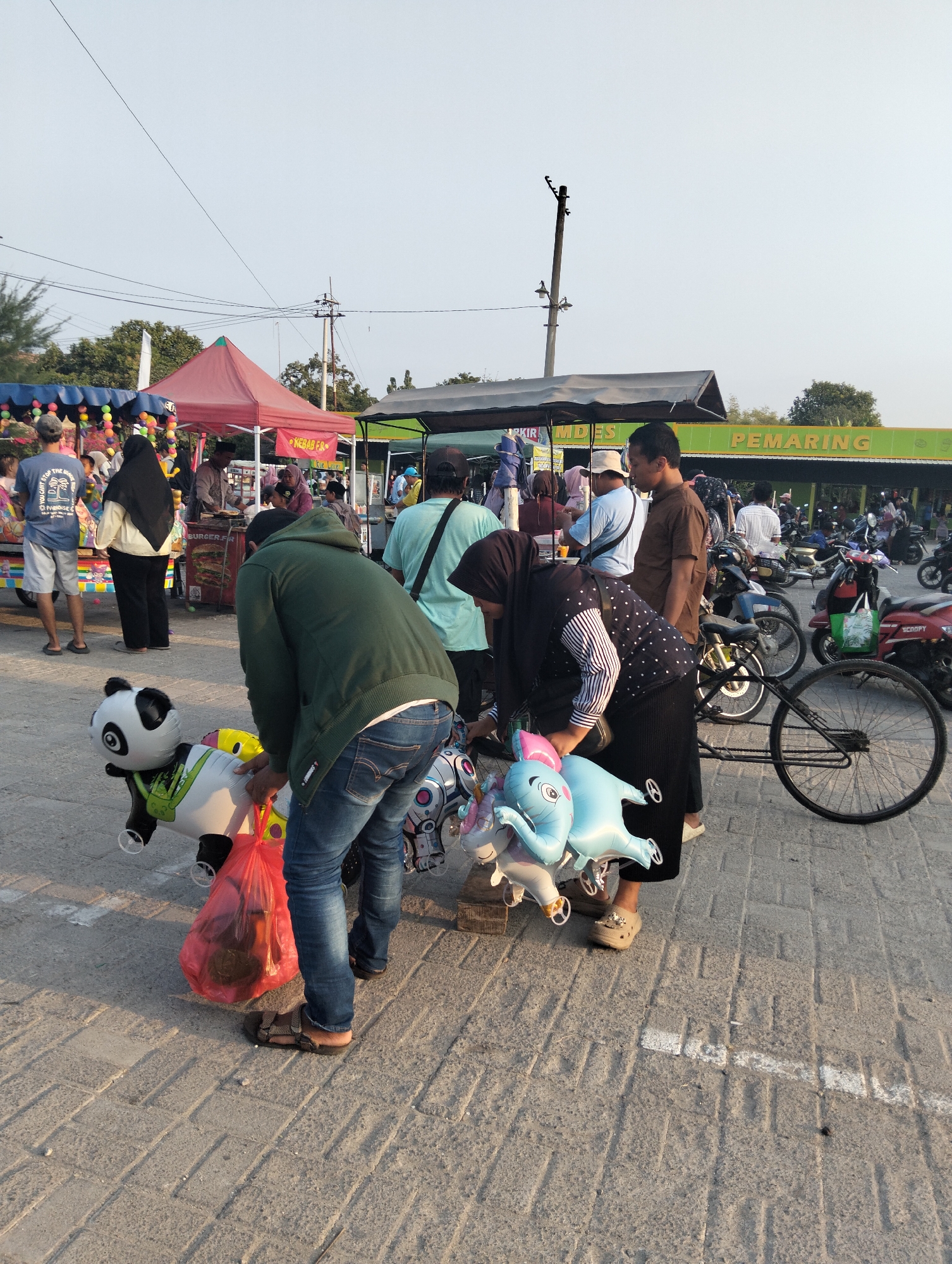 The image depicts an outdoor market scene in Drajat, Indonesia. In the foreground, a person wearing a green hooded jacket and blue jeans is tending to a collection of inflatable balloons, including a panda, an elephant, and a unicorn. The balloons are attached to a red plastic bag. To the left, a person in a blue t-shirt and shorts stands near a colorful cart filled with more balloons. In the background, several food stalls are visible, with awnings and signs advertising "BURGER.FR" and "PEMARING." A crowd of people are browsing the stalls and a row of motorcycles is parked to the right. The ground is paved with textured concrete blocks, and power lines are visible overhead. The overall atmosphere suggests a lively marketplace during daylight hours.