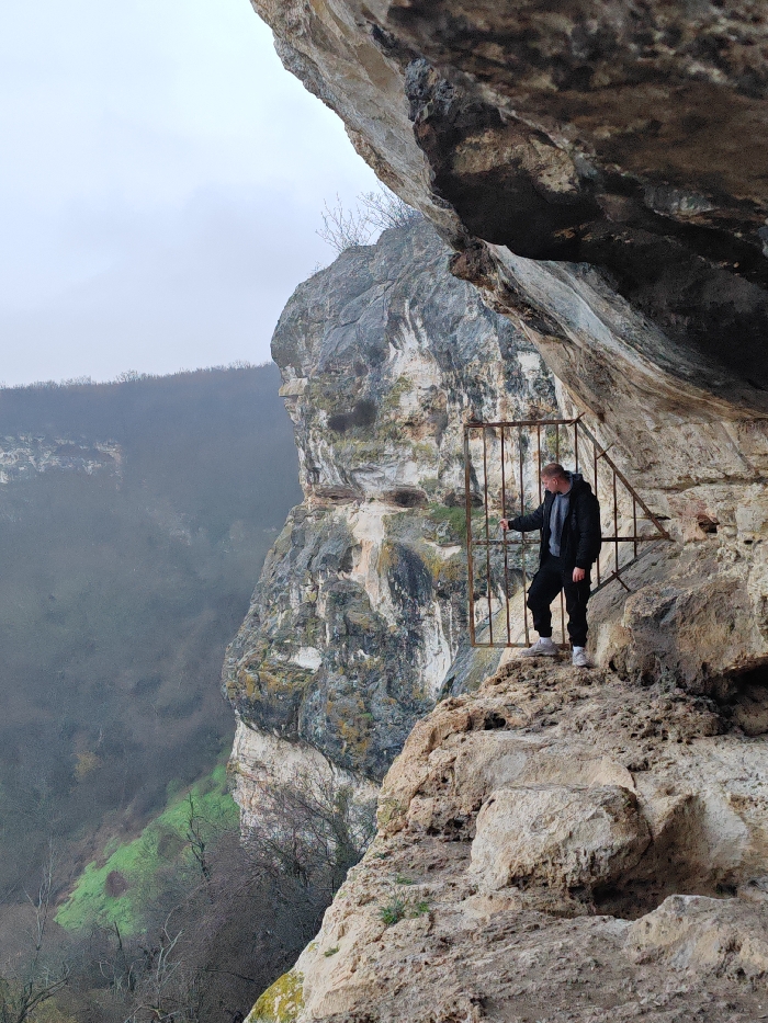 A single adult male is depicted standing on a rugged, uneven rock ledge on a cliff face, identified as part of the Shumen Plateau near the city of Shumen, Bulgaria. The individual, dressed in a dark hooded jacket, dark trousers, and light-colored athletic shoes, is positioned facing generally forward and slightly to his left. His right hand is grasping a vertical bar of a rusty, weathered metal gate. The gate consists of multiple vertical bars and horizontal crossbars, and appears to be integrated into the rock wall, potentially securing access to a cave or rock-cut dwelling. The male's head is turned slightly to his left, and his gaze is directed downward towards the expansive, mist-shrouded valley below, which features steep, tree-covered slopes with patches of green vegetation. The immediate foreground consists of a heavily textured, light-colored rock formation serving as the ledge. Above and to the right, a large rock overhang creates a partial ceiling. The sky is uniformly overcast and grey, indicating an absence of direct sunlight.