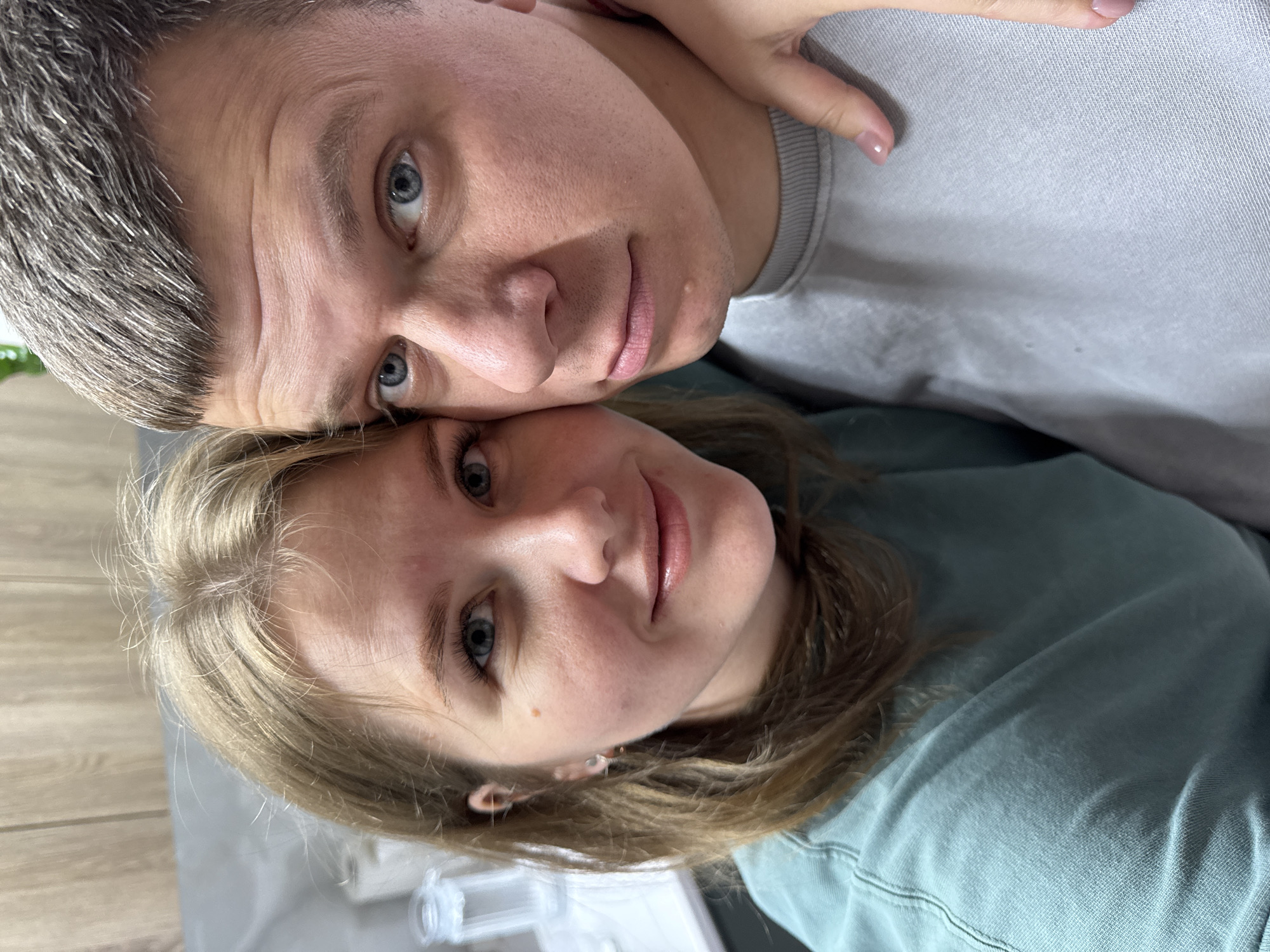 A close-up portrait features a man and a woman with their heads leaning together, both looking towards the camera. The man, positioned above and to the left, has short, salt-and-pepper hair and striking blue eyes. He is wearing a light grey crew-neck top. His expression is calm and direct.
The woman, to the right and slightly below the man, has long, light brown hair that frames her face and matches her blue eyes. She wears a muted teal or green long-sleeved top. A faint, soft smile plays on her lips, conveying a gentle and content demeanor. Her right hand, adorned with light pink manicured nails, is visible resting near the man's shoulder.
The background is softly blurred, indicating an indoor setting. A light-colored wooden surface can be discerned in the lower left corner, with other indistinct light-toned objects further in the background. The image, originating from Gornyi (Dvurechenskoe S/P), Russia, captures an intimate moment between the two subjects. There is no visible text, and the time of day is not explicitly indicated, though the lighting suggests an indoor environment.