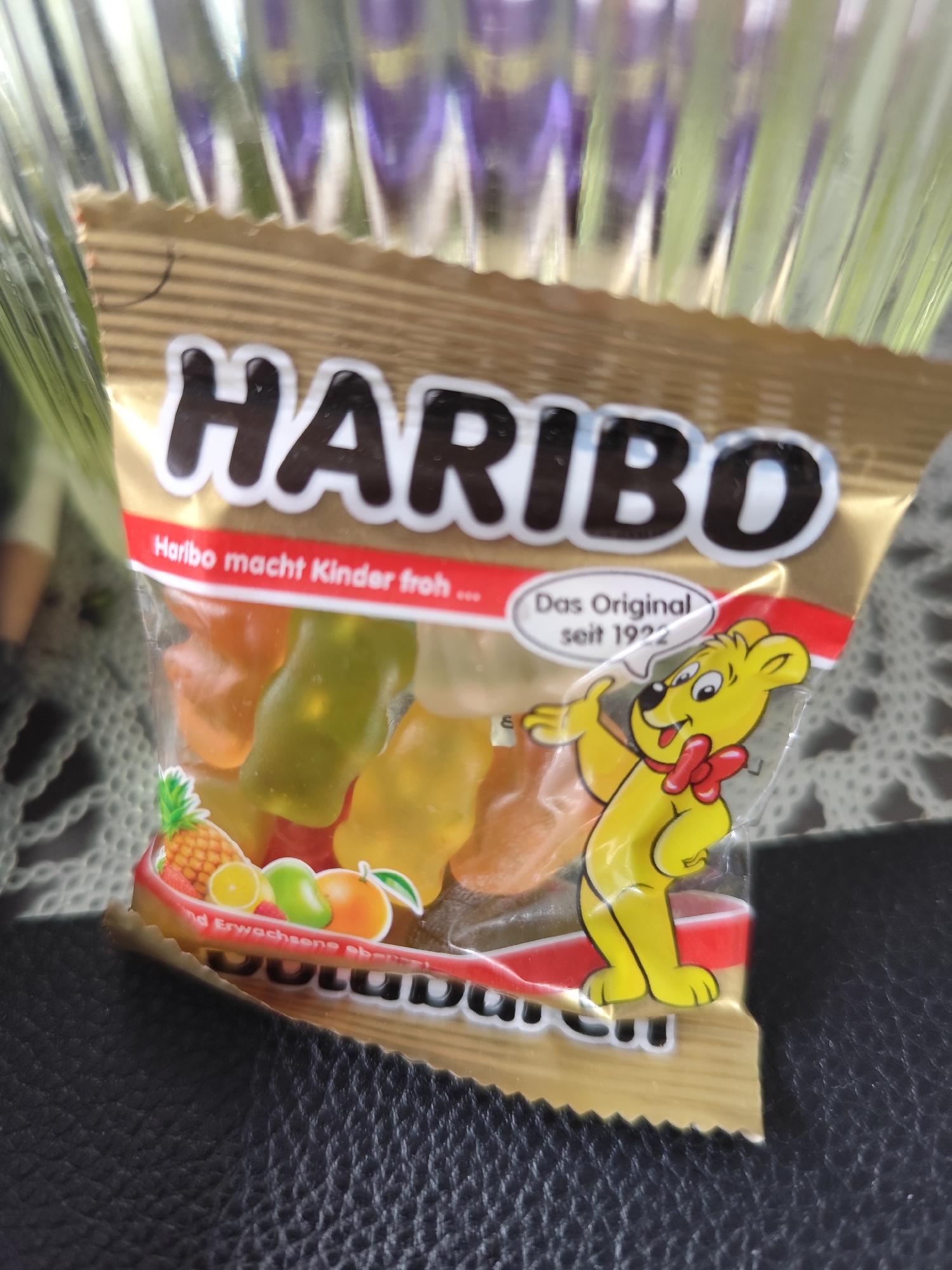 A bag of Haribo Goldbears candy is depicted in a close-up, indoor shot. The bag is partially open at the top, revealing the colorful gummy bears inside. The front of the bag features the Haribo logo, the slogan "Haribo macht Kinder froh..." (Haribo makes children happy...), and an image of the iconic Haribo bear character with a red bow tie. Below this, there is a graphic of various fruits including a pineapple, orange, and apple. The bottom of the bag has the words "GOLD...ERN" visible, suggesting it is a "Goldbären" (Goldbears) product. The bag itself appears to be made of a shiny, golden material. The background is blurred, showing vertical lines of light purple and green. The lower portion of the image shows a textured, dark surface, possibly leather. There are no people visible in the image. The image is well-lit, with the candy bag being the primary focus. The location in Dillingen an der Donau, Germany, is not visually evident in the provided image.