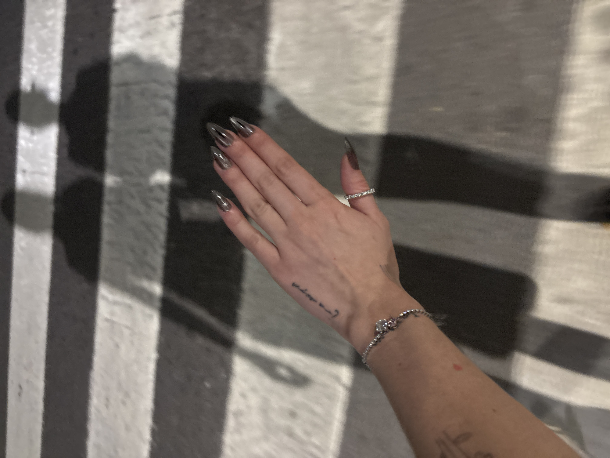 A person's left hand and forearm are visible in the image. The hand has long, stiletto-shaped nails painted in a metallic, transparent color. A ring adorns the ring finger, and a delicate bracelet is around the wrist. A small tattoo is seen on the inner forearm. The hand is placed on a surface marked with alternating dark and light stripes, reminiscent of a crosswalk. The location is presumed to be the city of Buenos Aires, Argentina.