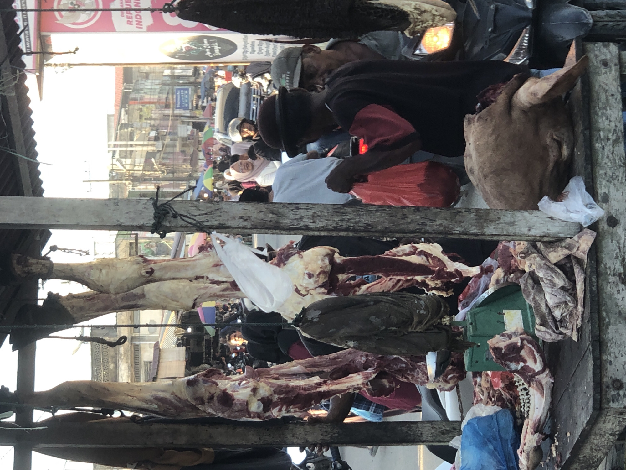 The image depicts a butcher shop in Kotapantonlabu, Indonesia. Hanged pieces of meat, including what seems to be a head, are displayed in the foreground. Several men are present; one is handling a red plastic bag, likely a customer. There's a scale on the wooden stand to weigh the meat. The background shows a busy street scene with buildings, signs (one with "REPUBLIK INDONESIA"), and other people.
