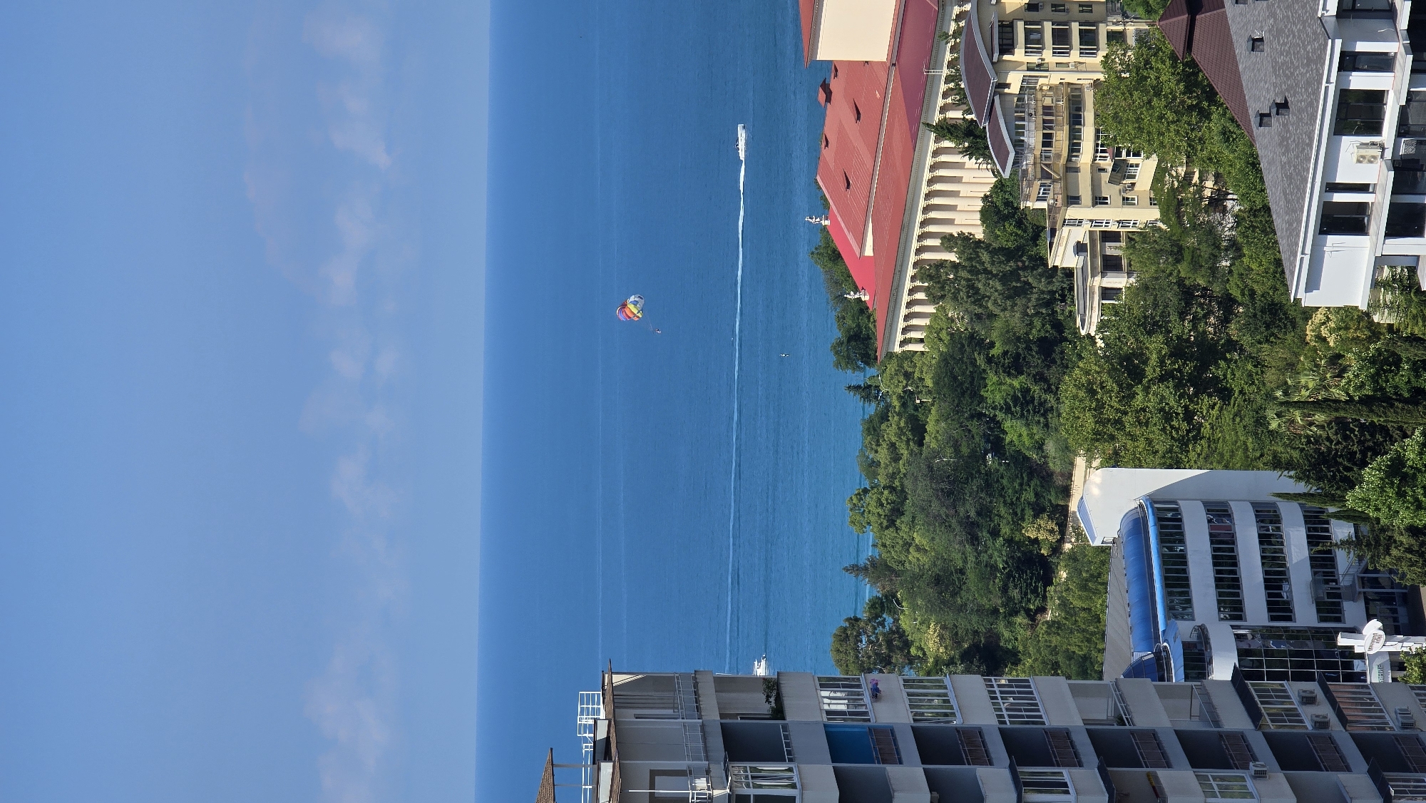 An aerial, vertical medium shot captures a bright, sunny day in Sochi, Russia, showcasing the city's coastline and architecture. The deep blue Black Sea fills the top two-thirds of the frame, with a thin line of pale clouds on the horizon separating it from a clear, light blue sky. A colorful parasail, carrying an unseen person, floats gently in the upper right quadrant of the sea. To the left of the parasail, a white speedboat leaves a distinct wake as it cruises across the water. Closer to shore, a much smaller white boat is visible.
The foreground is dominated by various buildings and lush greenery. On the far left, a tall, white apartment building with multiple balconies and blue accents juts into the frame. To its right and slightly further back, a curved building with a blue roof and large windows stands out. Behind these, a dense canopy of green trees covers the middle ground, interspersed with the red-tiled roofs and classical columns of a grand building. Yellow, modern apartment buildings with numerous balconies are nestled among the trees and on the slopes. In the bottom right corner, a contemporary building with a grey roof and large windows is partially visible. The overall impression is a scenic coastal city bathed in sunlight.