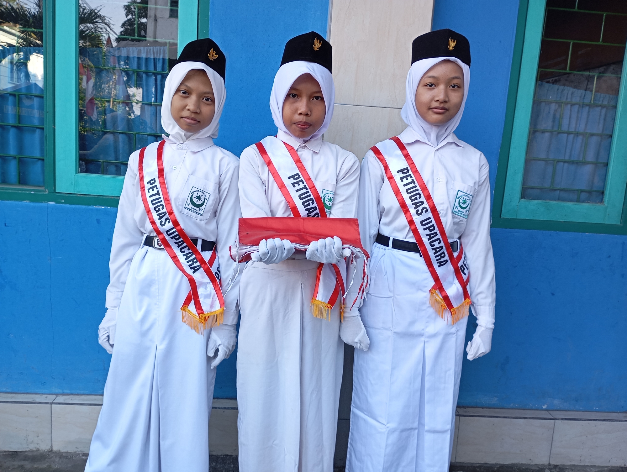 Three young individuals, likely students, stand in front of a blue wall. They are dressed in white uniforms, complete with white hijabs and black hats adorned with a gold emblem. Each person wears a white sash with red trim and gold fringe, bearing the red text "PETUGAS UPACARA" which translates to "Ceremony Officers." The person in the middle is holding a red object wrapped in white cloth. The environment suggests an outdoor setting, possibly near a building with windows, as indicated by the reflection on the left. The tiled ground and the blue painted wall are visible. The overall impression is one of formality and readiness for an event. Given the traditional attire and the uniform, it is likely that this scene is taking place in Mataram, Indonesia, where such student ceremonies are common.