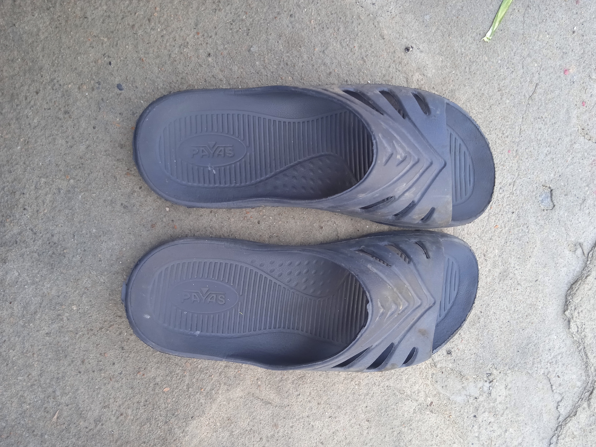 A pair of dark blue slide sandals are placed on a concrete surface. The sandals appear to be made of rubber or a similar material and have a textured footbed with the brand name "PAYAS" embossed in the heel area. Both sandals have a wide strap across the top with an angular, cut-out design. The concrete surface is rough and shows some signs of wear, with small pebbles and cracks visible. A thin, dry blade of grass is present in the upper right portion of the image. The lighting suggests an outdoor environment.