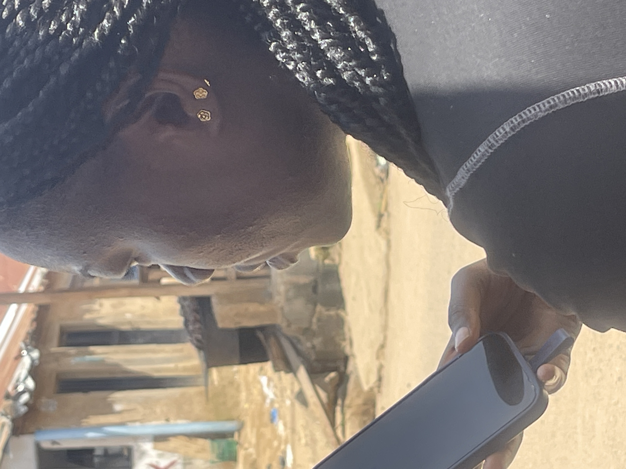 This image shows a close-up, sideways view of a person wearing braided hair and an earring. The person is holding a dark-colored smartphone. In the background, there is a rustic, outdoor scene with weathered wooden structures and what appears to be a dirt or sandy ground. The lighting suggests it might be daytime, with sunlight casting shadows. The overall impression is of an informal, possibly urban or semi-urban setting. There is no visible text in the image. The context provided indicates the location is Abuja, Nigeria.