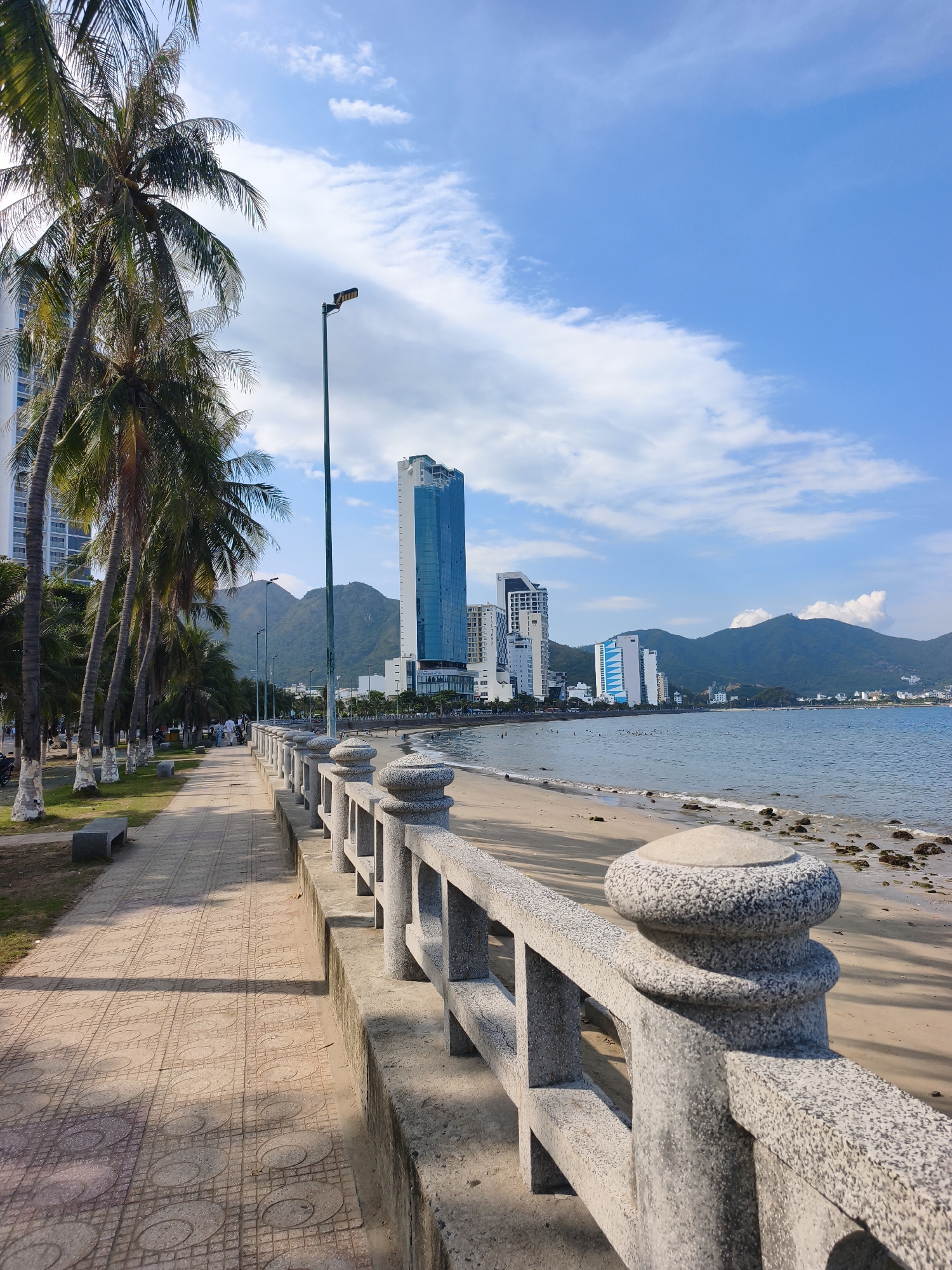 The image shows a scenic view of the beachfront in Nha Trang, Vietnam. A paved walkway runs alongside a sandy beach, bordered by a stone railing. Tall palm trees line the walkway, casting shadows on the patterned tiles. In the background, several modern high-rise buildings stand against a backdrop of lush green mountains. The ocean stretches out to the horizon under a bright blue sky with scattered white clouds. A few people can be seen walking on the path and swimming in the sea.
