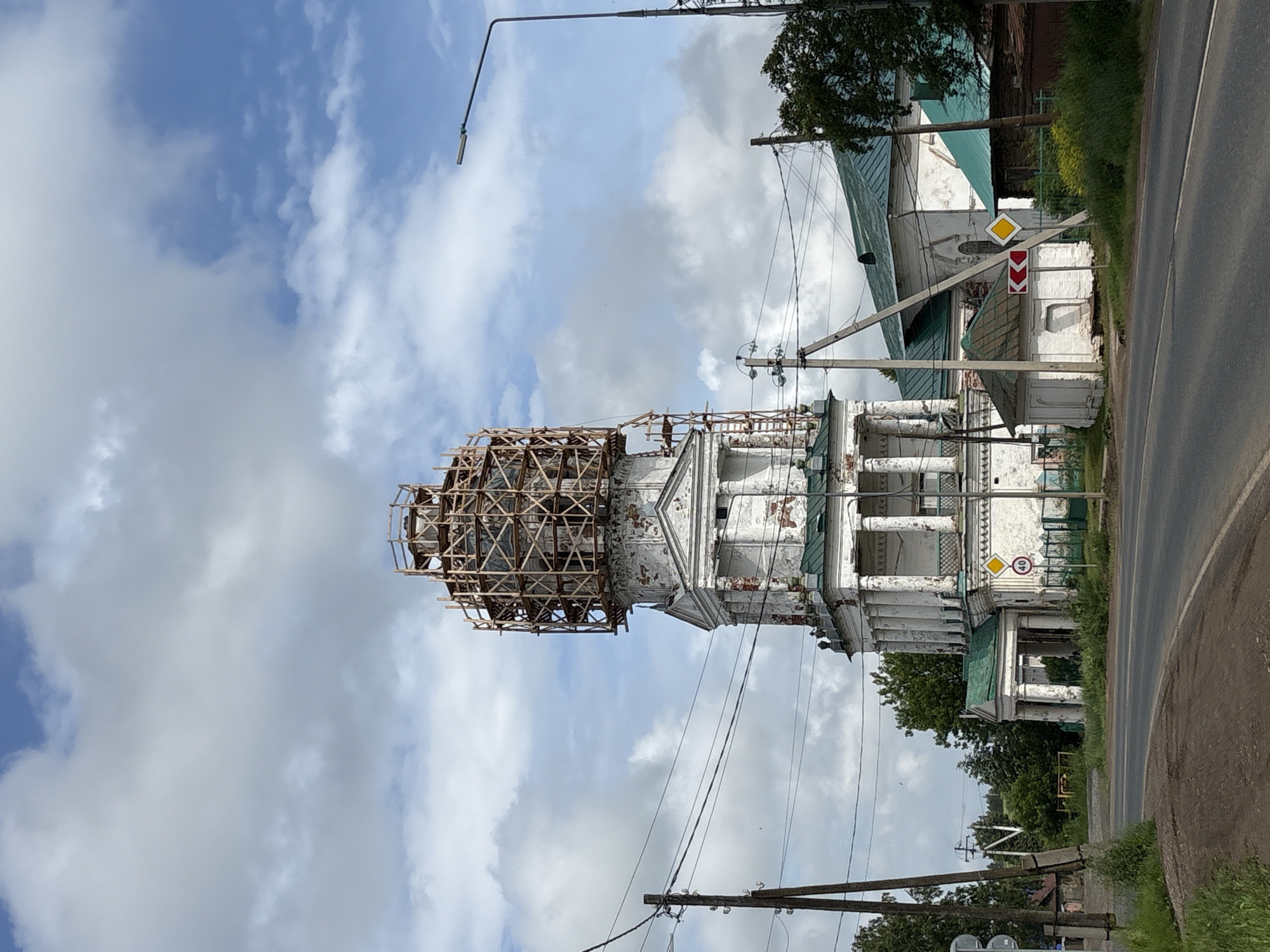 A tall, white, multi-tiered church structure is the central focus of the image, located in the village of Kstinino, Russia. The upper section of the church is enclosed by extensive wooden scaffolding, suggesting ongoing renovation or construction. The building exhibits signs of age, with peeling paint visible on its white facade and stone columns. Green roofs are present on adjacent structures. A paved road curves in the foreground, with grassy verges and some vegetation. Electrical poles and wires are visible throughout the scene. Traffic signs, including a speed limit sign (40) and directional signs, are present near the road. The sky is partly cloudy with a mix of blue sky and white clouds. No people are visible in the image.