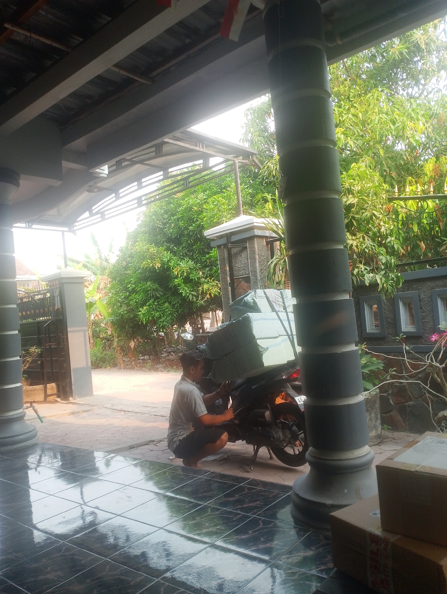 A man in a gray t-shirt and black shorts crouches next to a motorcycle loaded with large packages wrapped in light green plastic. The motorcycle is parked on a paved area in front of a building with a tiled floor and a covered porch. The porch is supported by thick, cylindrical pillars. In the background, there is a gate, more foliage, and a stone wall with decorative rectangular openings. The scene appears to be outdoors in a residential or commercial area, likely in the city of Kebonsari, Indonesia, given the context.