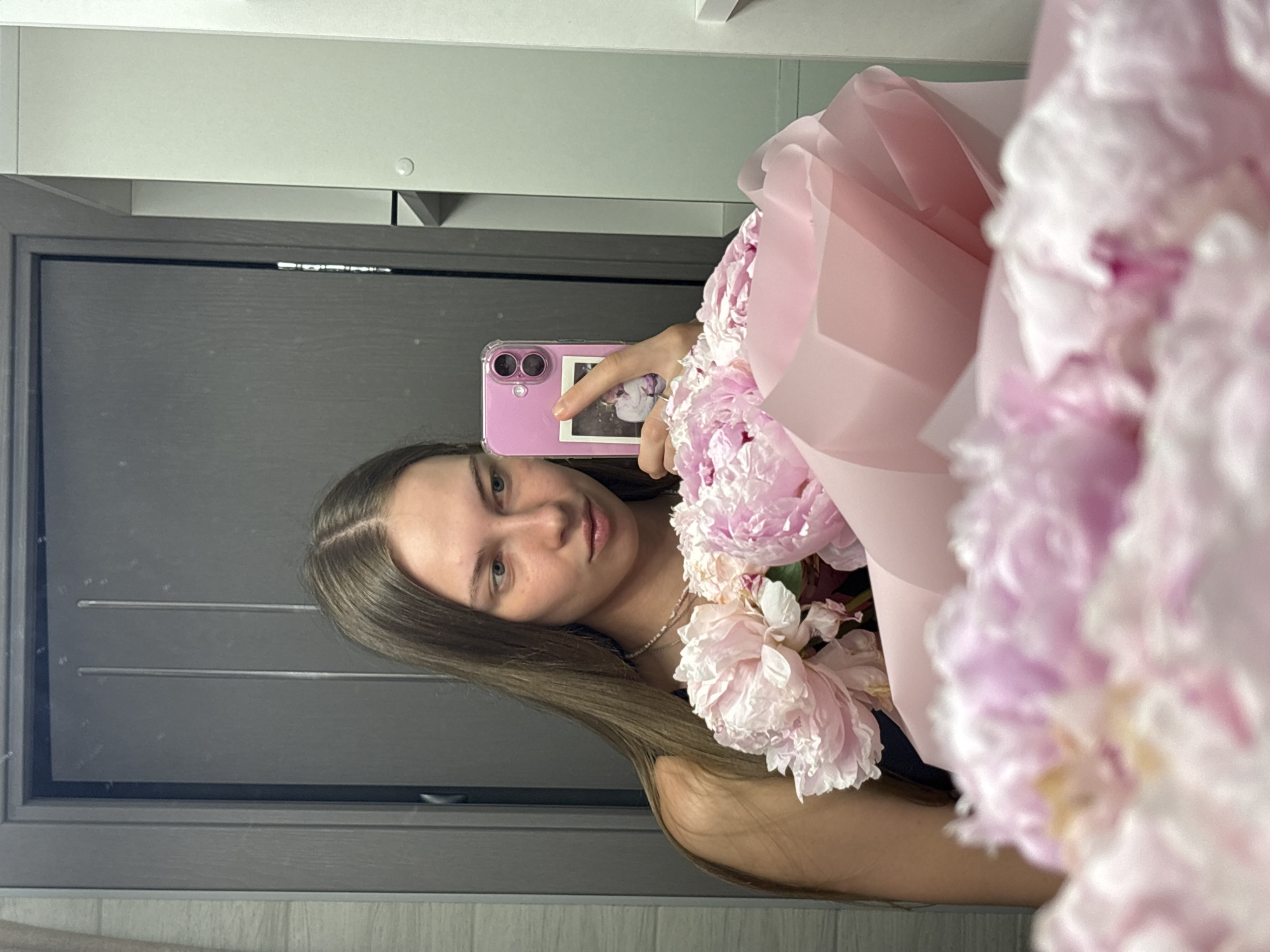 A young woman with long, straight brown hair takes a selfie in a mirror, holding a bouquet of pink peonies. She is wearing a dark top and a beaded necklace. Her phone, with a pink case and a small picture adhered to it, is visible in her right hand. The background appears to be an indoor setting with a door and part of a closet visible in the reflection. The overall ambiance is intimate and personal.
This image could have been taken in an apartment in the city of Perm, Russia.