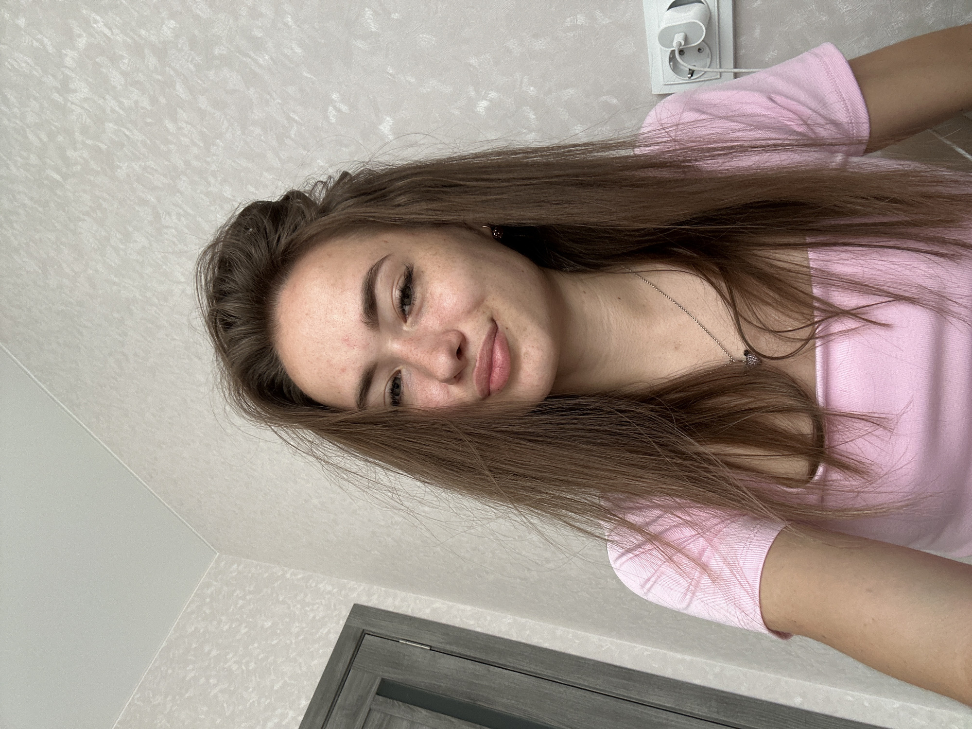A young woman with brown hair, fair skin, and freckles poses for a selfie in an indoor setting. She is wearing a pink t-shirt and a silver necklace with a heart-shaped pendant. Her hair is partially tied up, with loose strands framing her face. The background shows a textured wallpaper and a door on the left side of the frame, with an electrical outlet on the right. The lighting suggests an indoor, possibly residential environment, given the presence of a door and an electrical outlet.