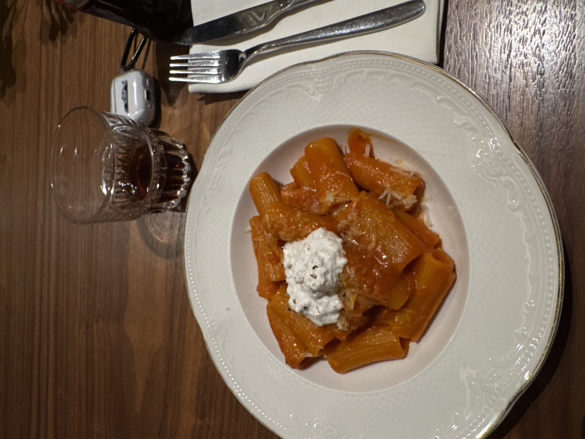The image shows a plate of pasta with tomato sauce and ricotta cheese, placed on a dark wooden table. Next to the plate, there is a glass of dark liquid (likely a drink), a butter knife, and a fork on a white napkin. The plate is white with a decorative edge. The pasta is a tube-shaped variety. There is no evidence to confirm the location as Yerevan, Armenia.