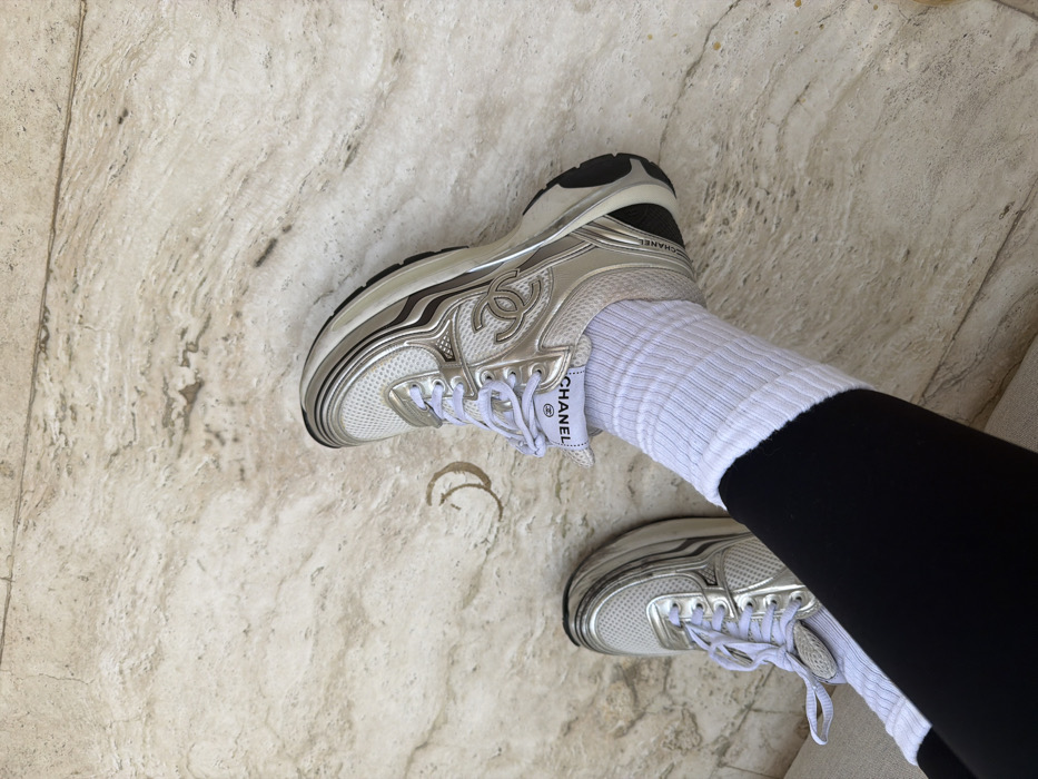 A close-up, top-down view depicts a person's lower legs and feet, clad in black trousers, white ribbed socks, and white and silver Chanel sneakers. The right sneaker prominently displays the Chanel 'CC' logo on the midfoot and 'CHANEL' branding on the tongue and heel. The footwear consists of white mesh and silver metallic overlays. The feet rest on a light-colored, polished marble or stone floor featuring natural veining and a faint, crescent-shaped light brown stain. This environment, characterized by luxury apparel and high-end flooring, is consistent with upscale residential or commercial interiors found throughout Dubai, United Arab Emirates. A specific address or landmark within Dubai is not discernible from the image.