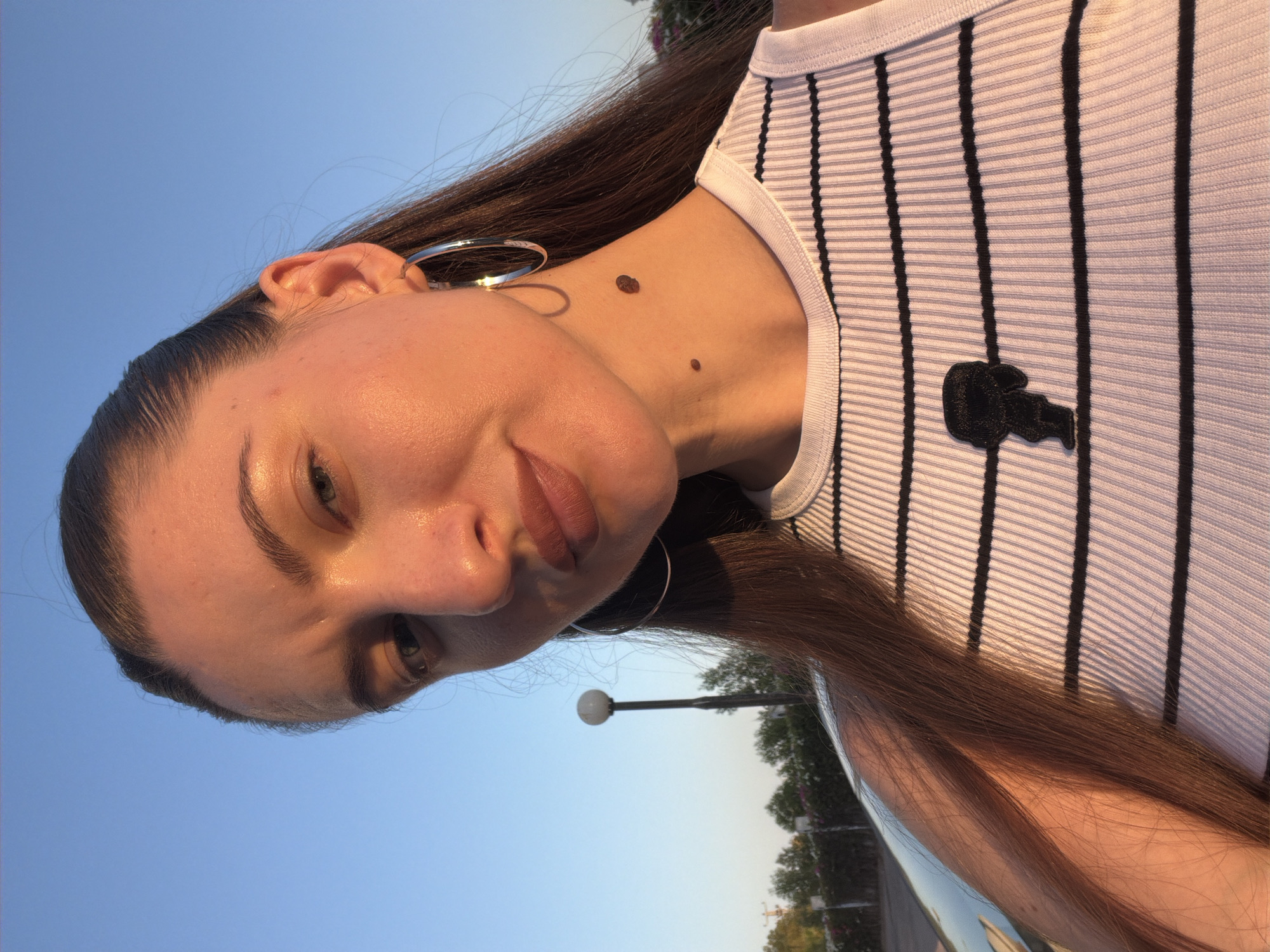 In Sharm El Sheikh, Egypt, a young woman with long brown hair, wearing a white ribbed tank top with black stripes and a black graphic, poses for a selfie outdoors. She has a mole on her neck and wears hoop earrings. The setting sun casts a warm glow on her face and skin. In the background, a street lamp and trees are visible.