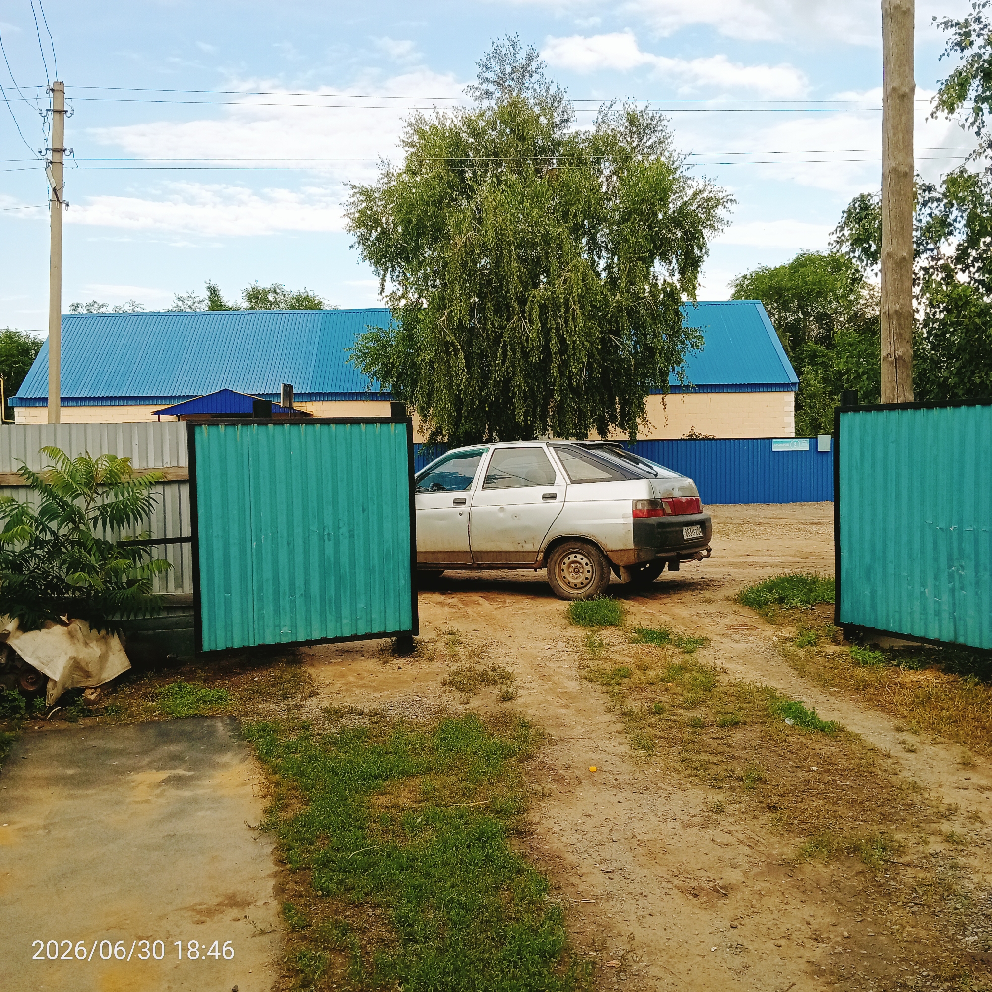 The image shows an outdoor scene in what appears to be a rural or semi-rural setting. The main subjects are a silver sedan parked on a dirt path and a large building with a bright blue corrugated metal roof. In the foreground, there are two gates made of turquoise corrugated metal, one of which is partially open. A chain-link fence is visible behind the turquoise gate on the left.
A large, leafy tree with light green foliage dominates the middle ground, partially obscuring the blue-roofed building. The building itself has lighter colored walls and appears to be a workshop or storage facility. There are power lines and poles visible against a cloudy blue sky, suggesting the presence of electricity in the area.
The ground is a mixture of dirt, gravel, and patches of grass. The car is parked at an angle, facing away from the viewer and towards the building. It appears to be a somewhat older model, with some signs of wear and tear, including rust on the lower body panels.
The lighting suggests it is daytime, possibly late afternoon given the angle of the shadows, though it's difficult to be certain. The weather appears to be partly cloudy. A timestamp is visible in the bottom left corner of the image, reading "2026/06/30 18:46". There is no discernible activity taking place; the scene appears to be static. The overall impression is one of a utilitarian or industrial outdoor space, perhaps a yard or driveway.