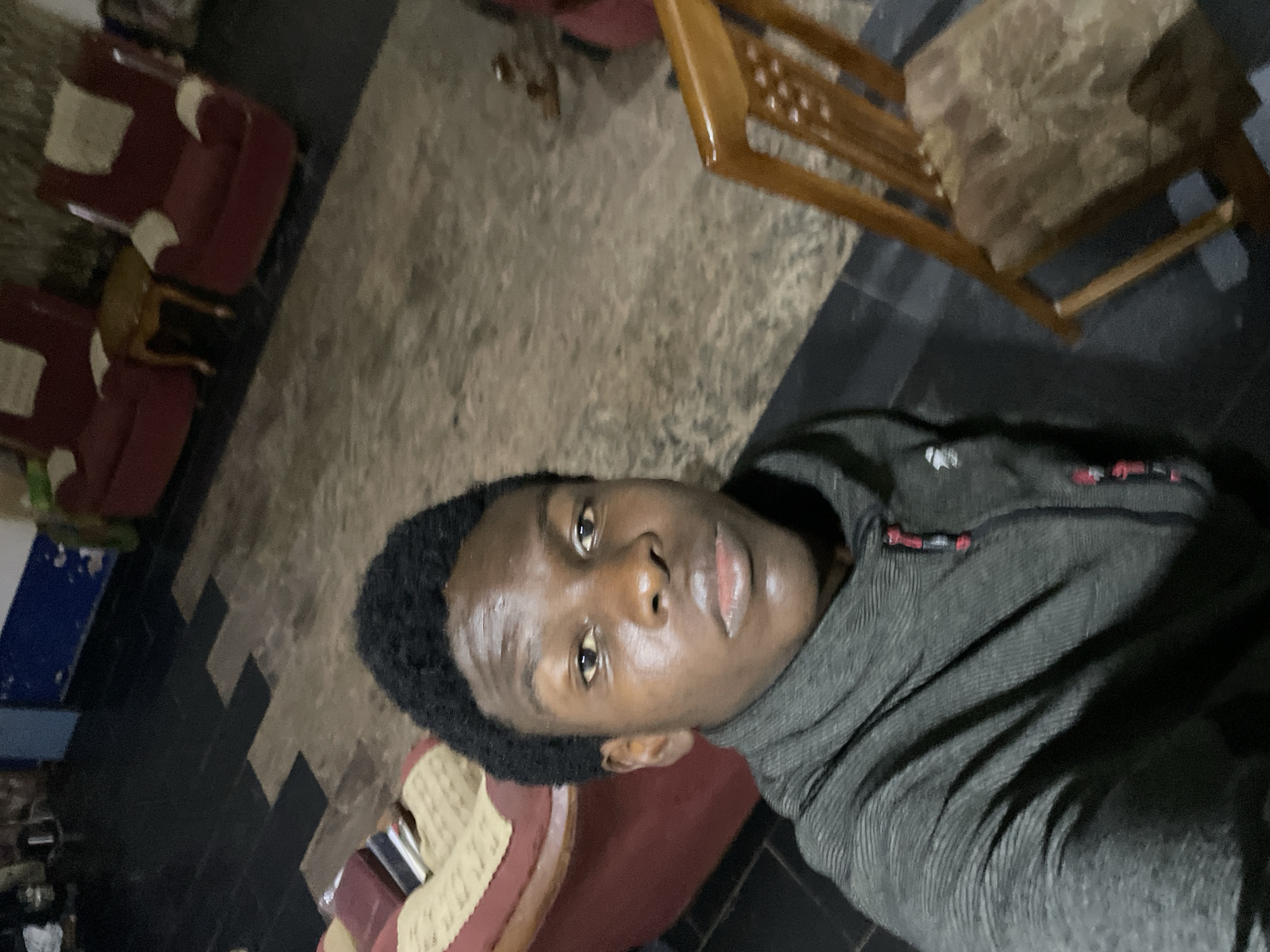 A close-up selfie of a young dark-skinned man with short black hair, wearing a dark gray hooded shirt. The background shows part of a room with a tile floor, multiple chairs (wooden and upholstered), and a small table. The location is Rafin Bauna, Nigeria.