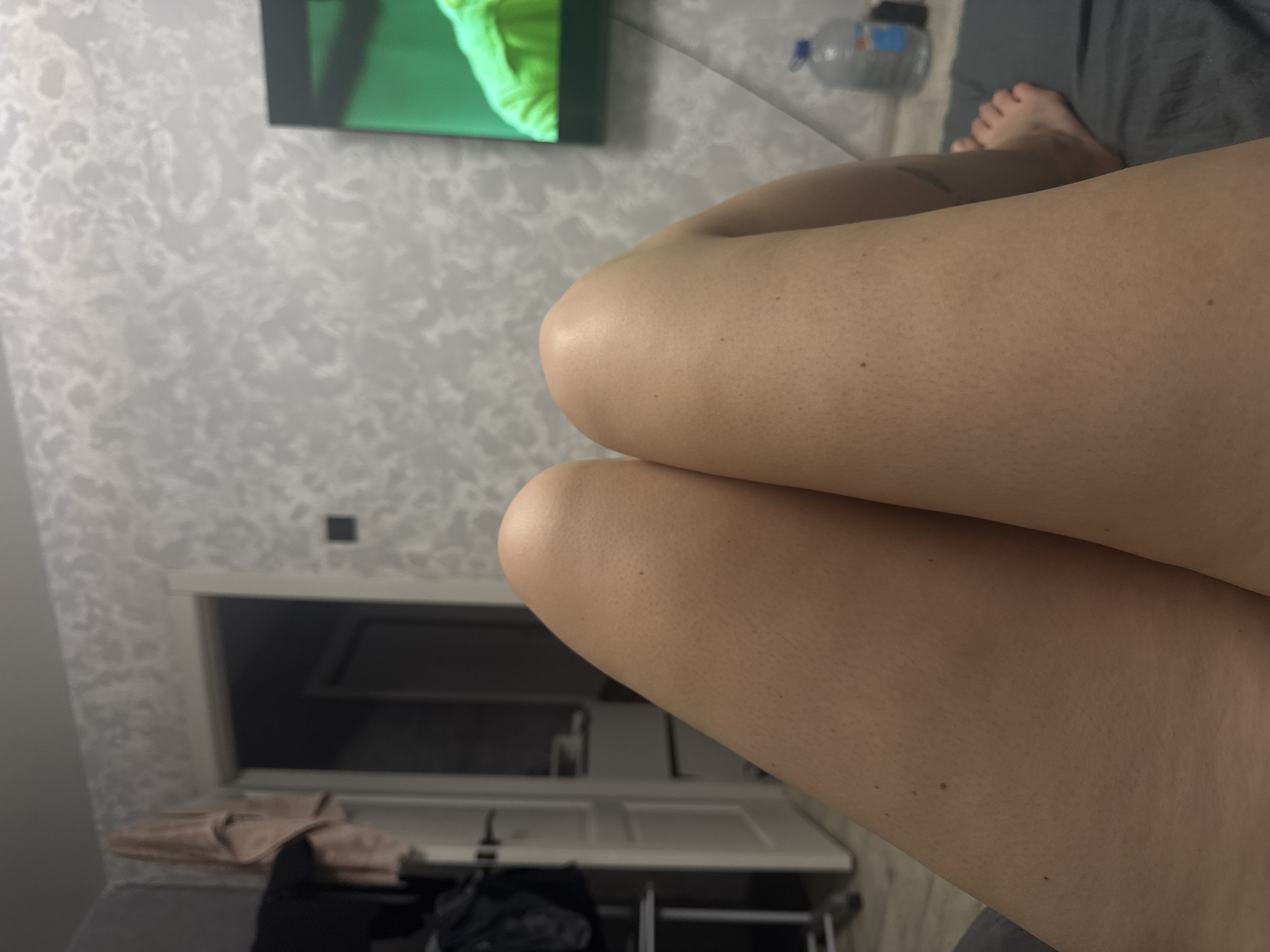 A first-person view captures the observer's bare legs, positioned centrally, with a domestic interior in the background. The legs appear light brown or tan, displaying visible skin texture and minor blemishes. The right foot is partially visible in the lower right, resting on a dark grey fabric surface, likely a bed or sofa.
The background wall is covered with light grey wallpaper featuring a subtle, embossed floral or abstract pattern. To the left, a white doorway leads into a dimly lit area, with a white door partially open. A light pink fabric item is draped over the wall above the door frame. Below it, a dark garment is seen hanging on a light-colored structure, possibly a chair or stand.
To the right, a flat-screen television is mounted on the wall, displaying a bright green image, possibly of a person in green attire. A dark cable descends from the TV. A large clear plastic water bottle with a blue cap and label rests on the dark grey fabric surface in the lower right corner, near the television. The scene suggests an indoor setting, likely a bedroom or living area, in the city of Unknown, Unknown.