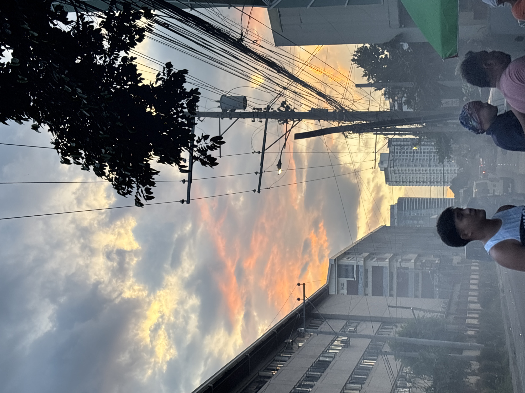 The image captures a street scene in Davao City, Philippines, during what appears to be dusk. The sky is filled with dramatic clouds, painted with hues of orange, yellow, and gray, suggesting either a sunset or sunrise. In the foreground, three individuals are partially visible. The person on the far left is a young male wearing a white tank top with black trim. Next to him, a person wearing a dark shirt and a patterned head covering is visible from the side. To the right, another individual in a pink shirt is seen from the side.
The urban environment is characterized by a dense network of electrical wires and poles that dominate the midground and background. Tall buildings, some appearing to be residential or office complexes with numerous windows, line the street. One building on the left has a long facade with rows of air conditioning units. In the distance, several high-rise buildings are silhouetted against the sky. Smoke or haze is present, particularly in the lower portion of the image, obscuring some details of the street and vehicles. A green canopy, possibly part of a street stall or vendor, is visible on the right side of the frame. The overall impression is a bustling city environment under a colorful, cloudy sky.